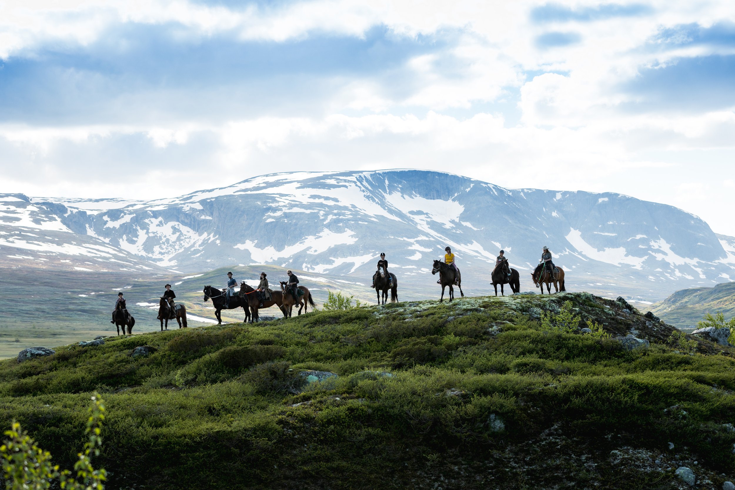 Ni ryttere på hesteryggen krysser en fjellrygg med snødekte fjell i bakgrunnen.