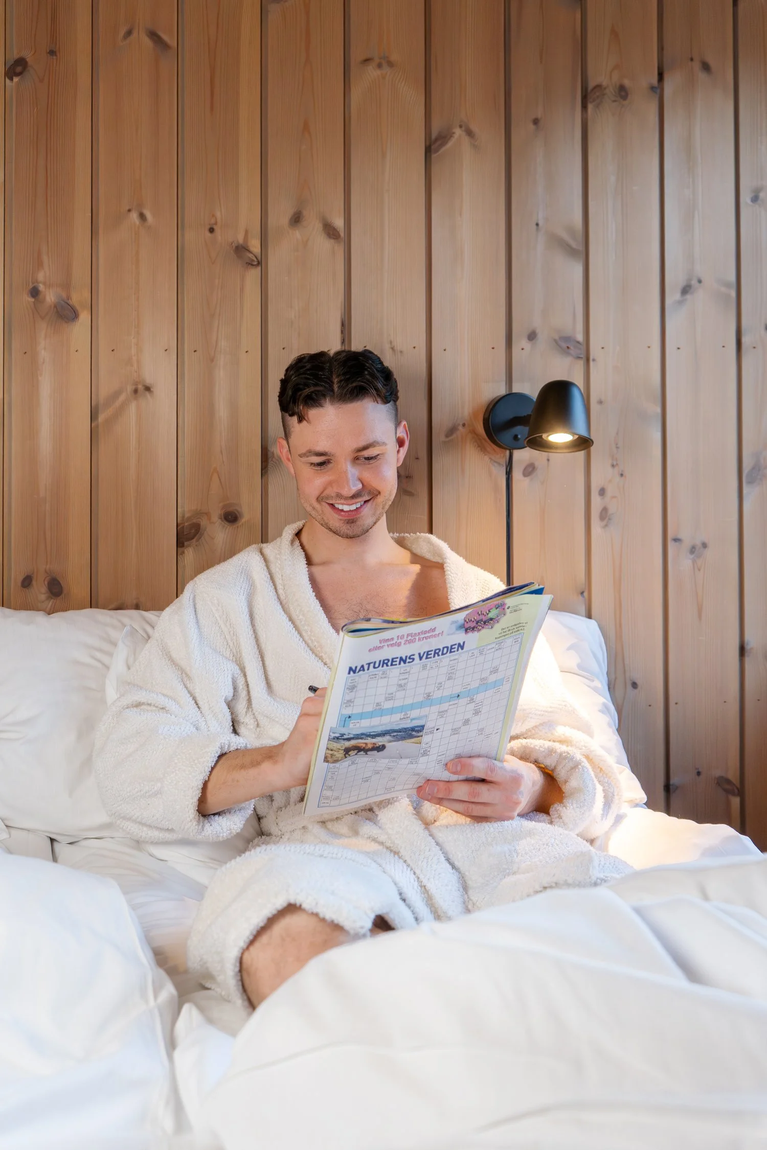 Man sitting in a bathrobe in bed solving crossword puzzles.