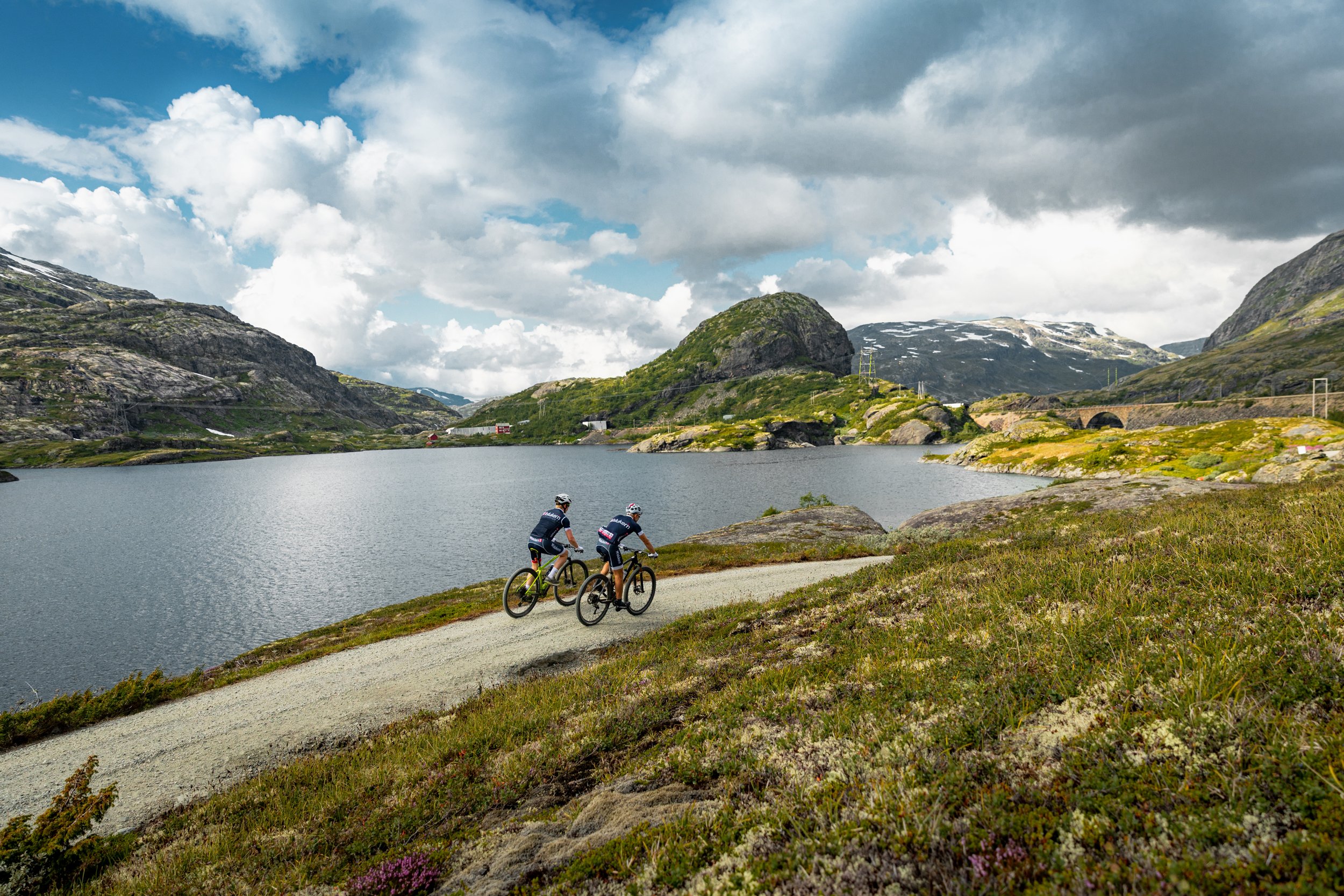 To personer sykler på en grusvei ved en innsjø omgitt av fjell med snø, under en skyet himmel.