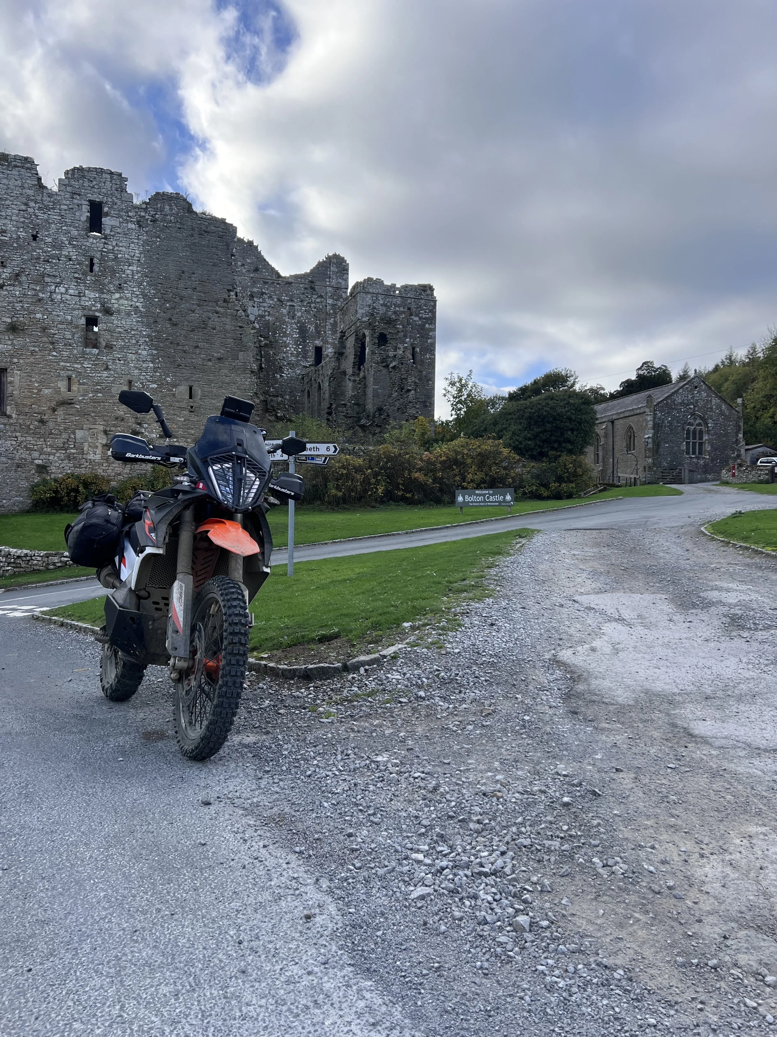Motorcycle parked on a gravel driveway in front of Bolton Castle ruins with a sign pointing towards the castle. The castle is an old stone structure situated on a grassy area with a cloudy sky overhead.