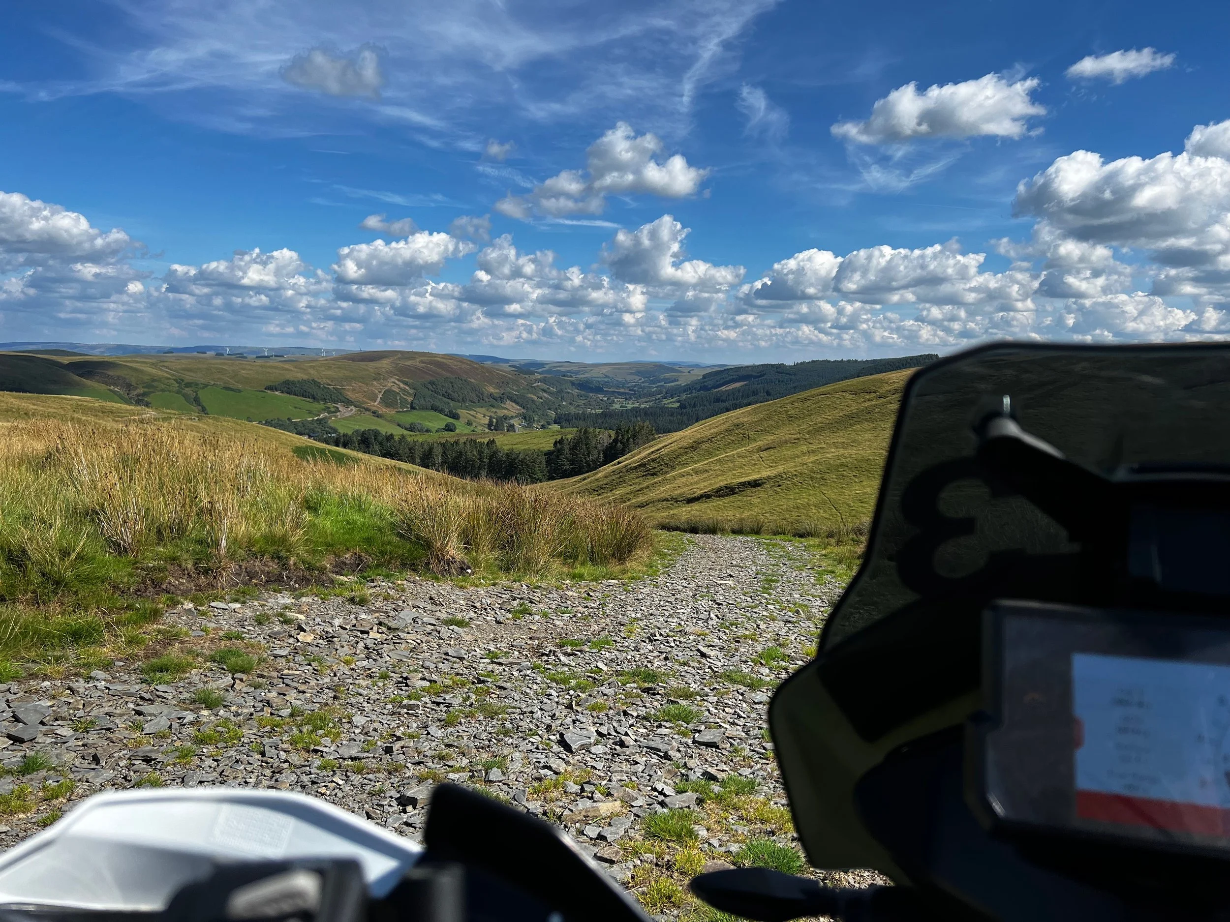 View of rolling green hills on a sunny day with cloudy sky, taken from a mountain biking trail, with parts of motorcycle helmets in the foreground.