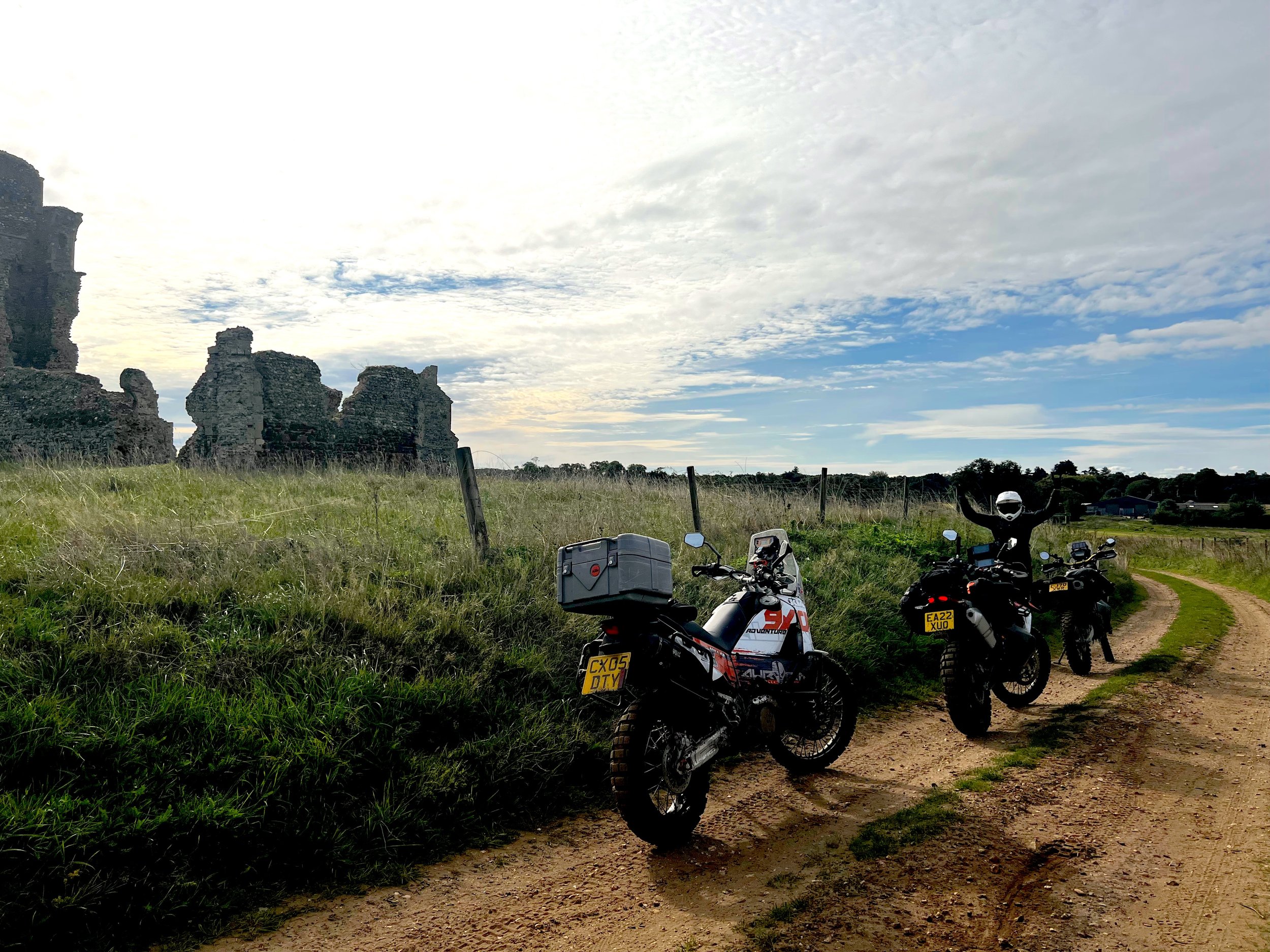 Three adventure motorcycles parked on a dirt path with an open field and ancient ruins in the background under a partly cloudy sky.
