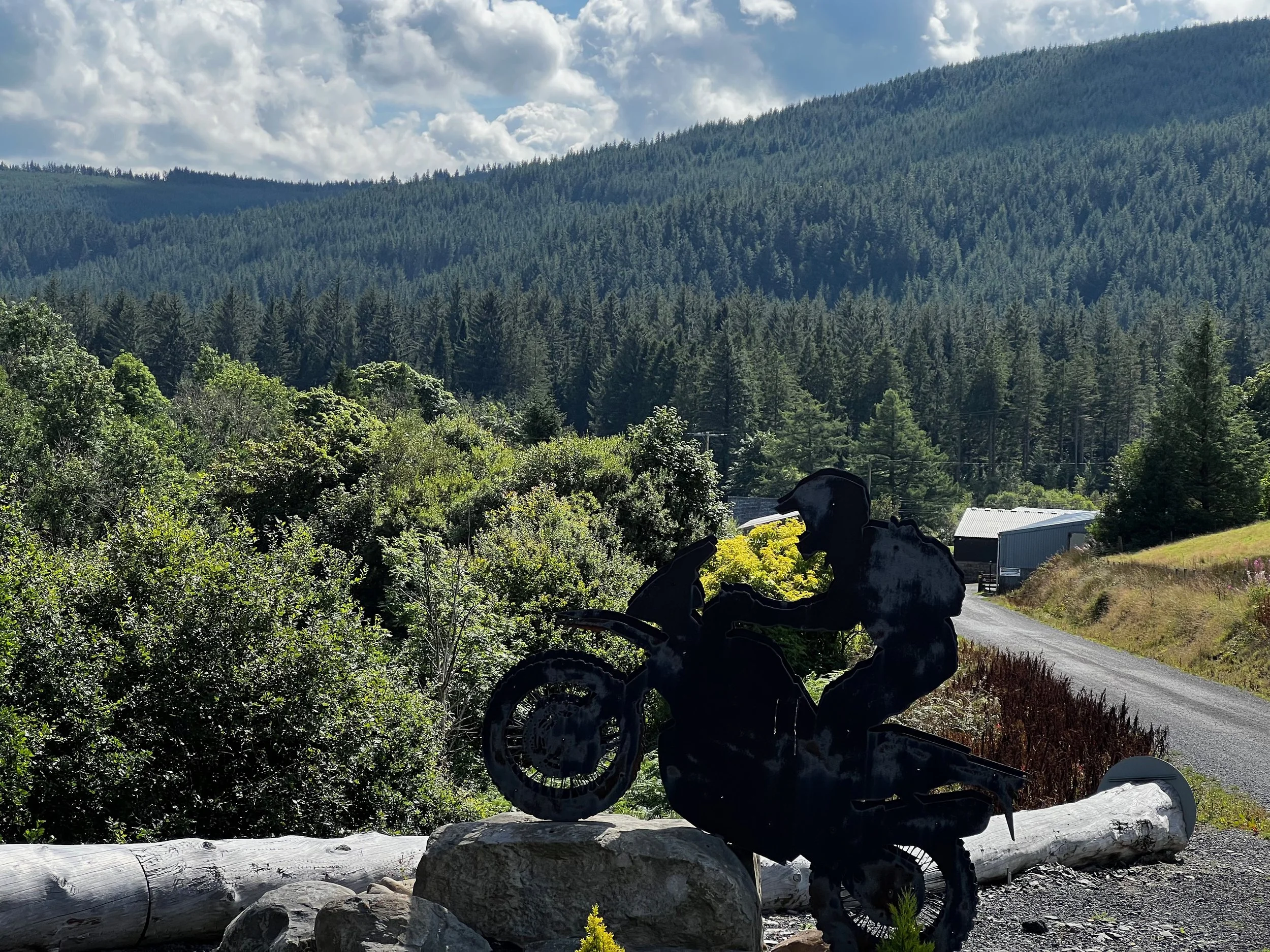 Metal silhouette sculpture of a motorcyclist riding on a motorcycle, placed on a stone along a rural road with lush green trees and mountains in the background on a partly cloudy day.