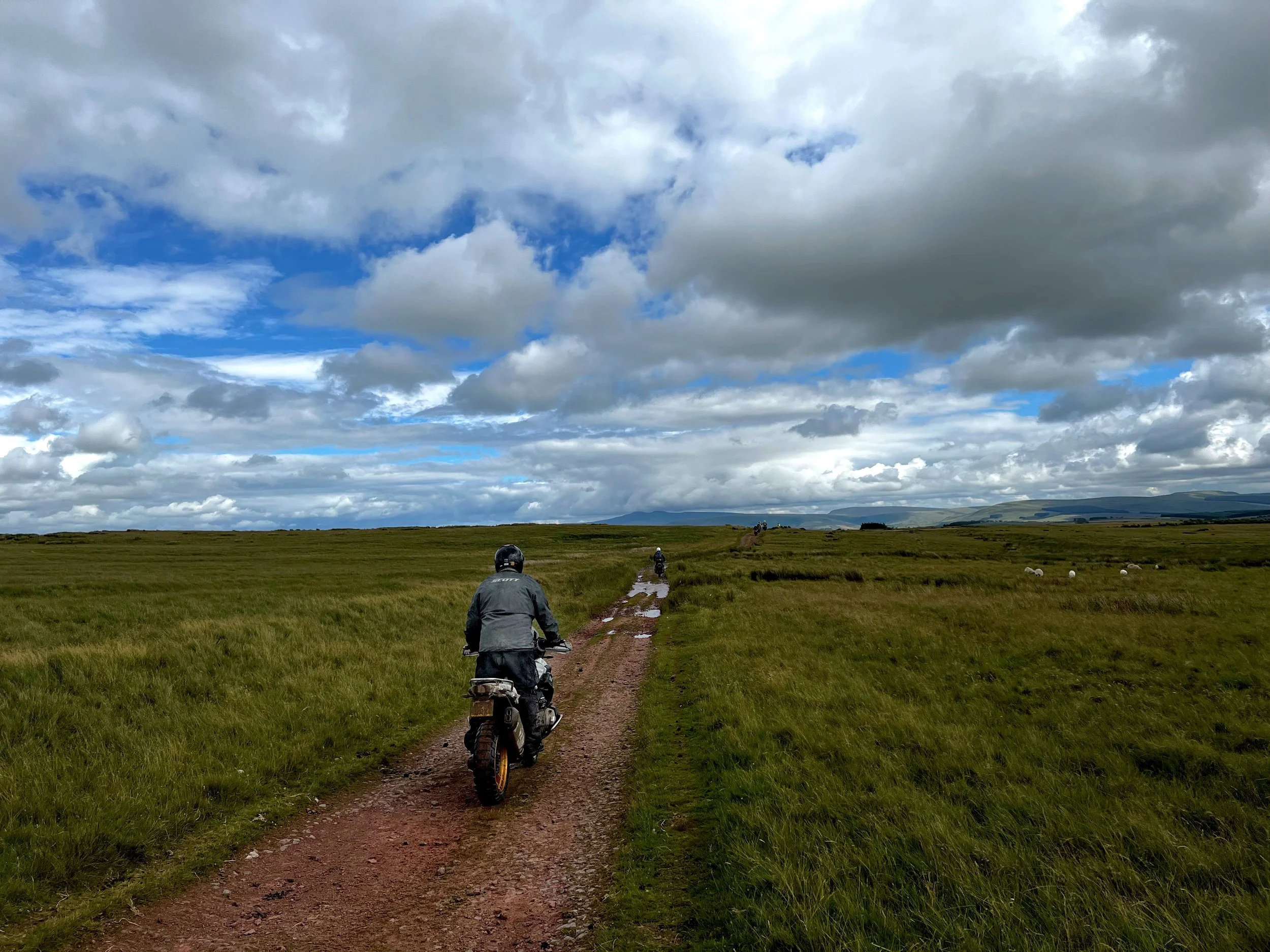 Two motorcyclists riding on a dirt path through a grassy field under a cloudy sky.
