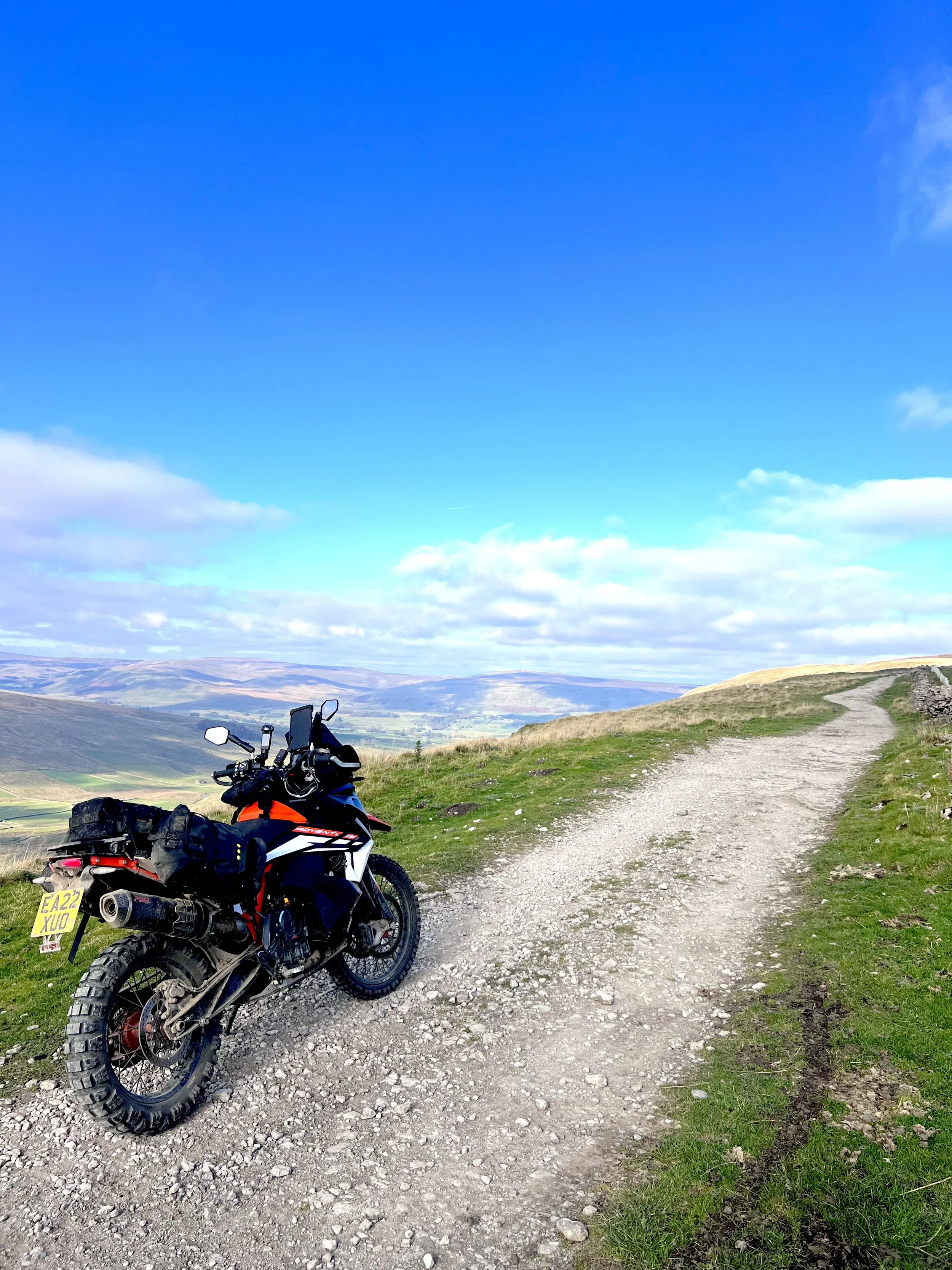 A black and orange motorcycle parked on a dirt trail in a hilly landscape with green grass, distant hills, and a partly cloudy sky.