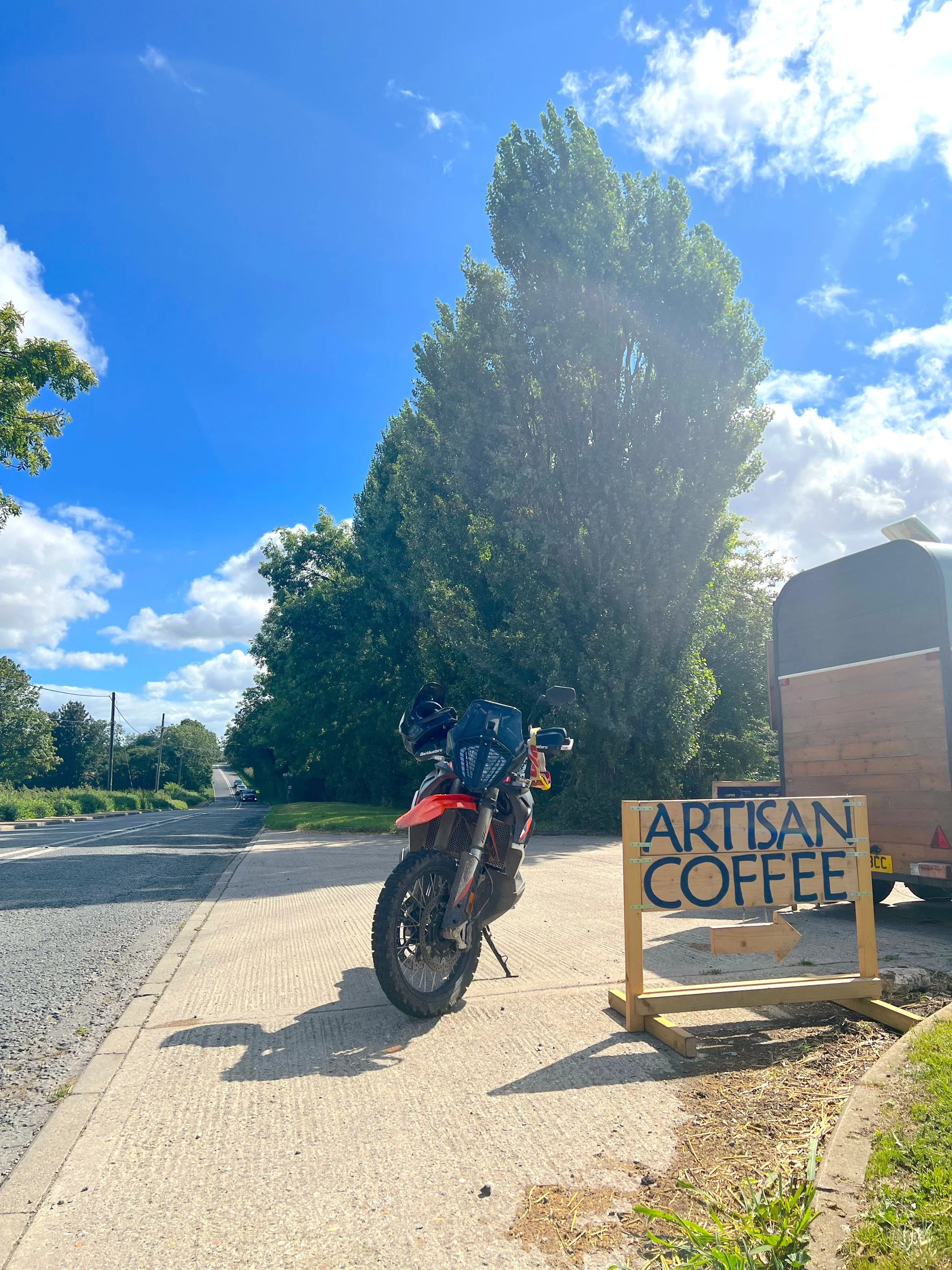 A motorcycle parked next to a wooden sign that reads 'Artisan Coffee' with an arrow pointing to the right, alongside a food trailer on the sidewalk under a bright blue sky with some clouds, tall trees in the background.