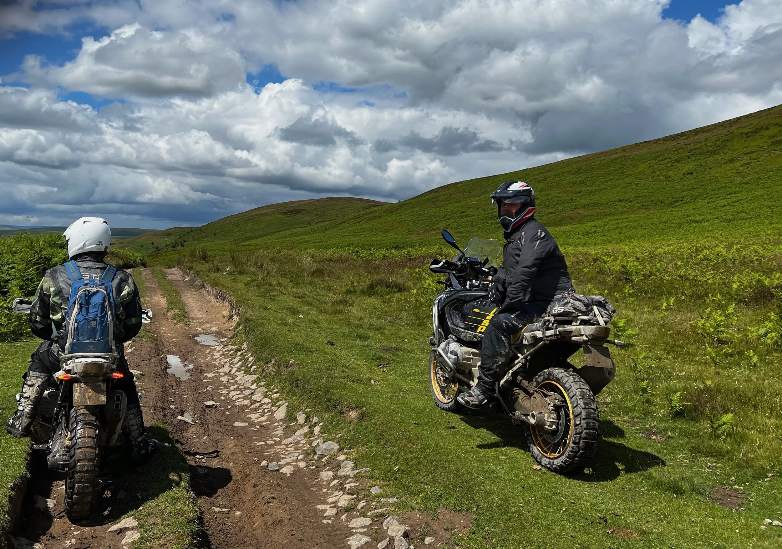 Two motorcyclists in black gear and helmets on a dirt trail in green hilly landscape with cloudy sky.