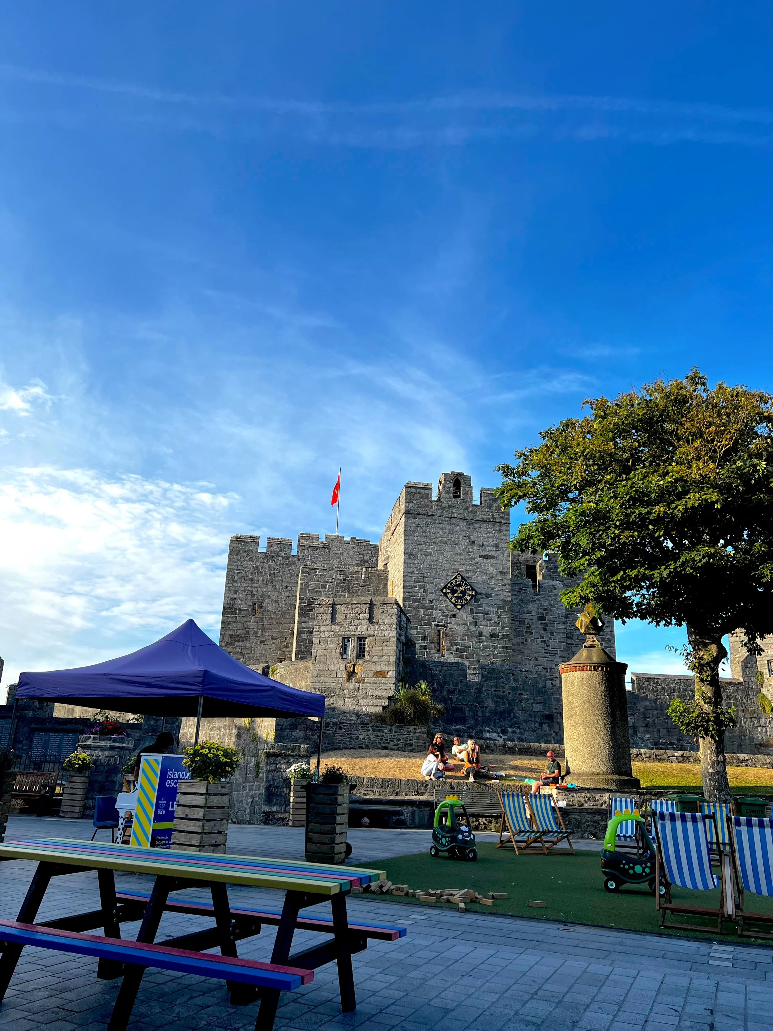 A stone castle with a flag on top, a clock, and turrets, situated on a hill. In the foreground, there is a tent, benches, lounge chairs, and children playing, with a tree on the right and a bright blue sky overhead.