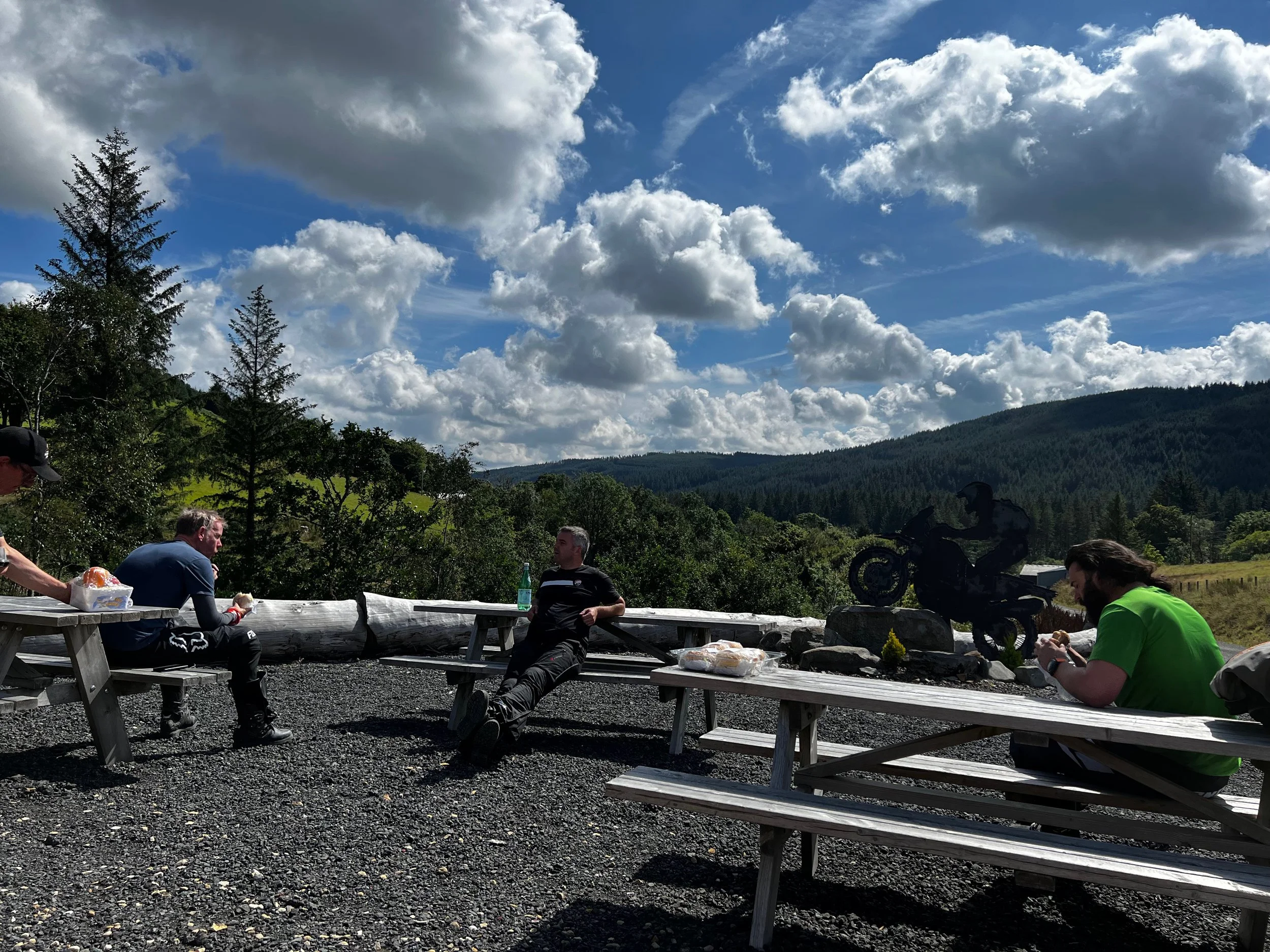 Four people sitting and relaxing outdoors at wooden picnic tables with a scenic mountain and forest background under partly cloudy sky.