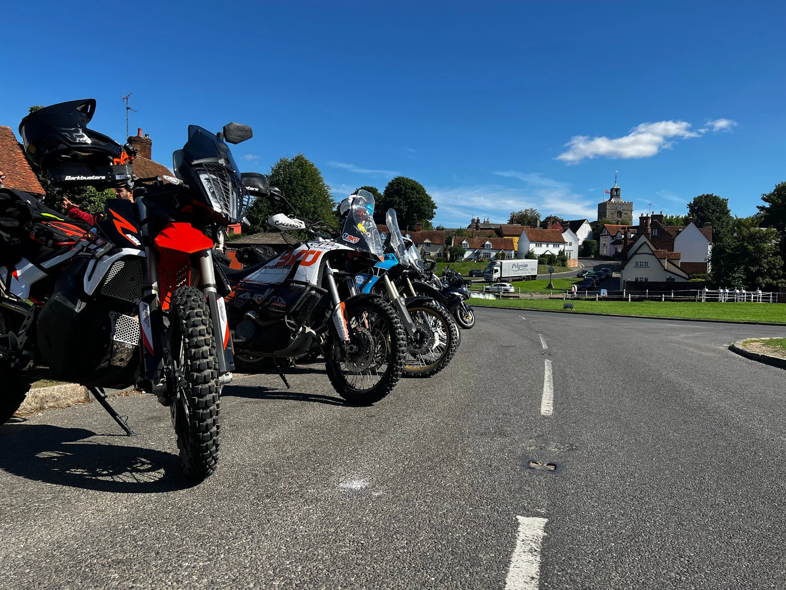A row of motorcycles parked on a street with a small town and green landscape in the background under a clear blue sky.