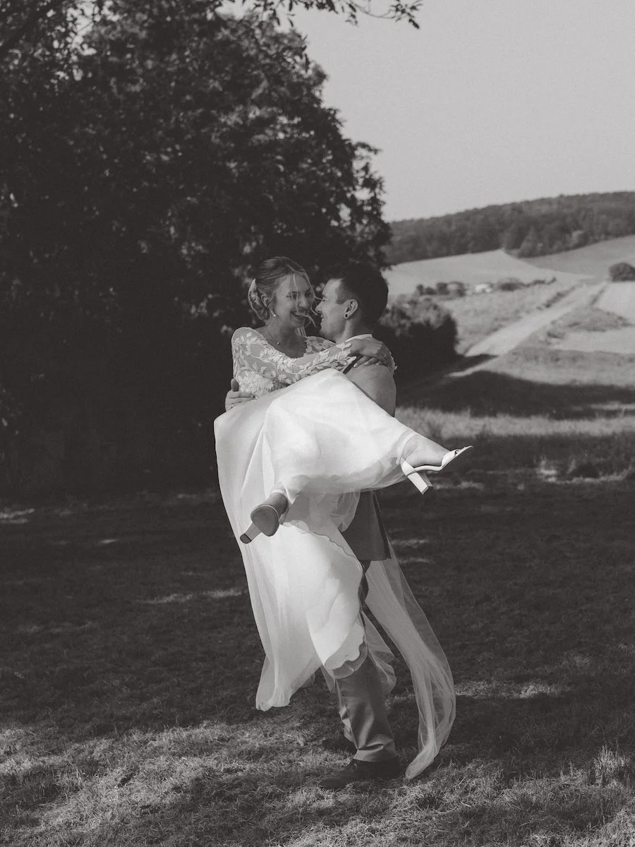 Der Bräutigam hält stolz seine Braut, die ausgelassen lacht, während eines Hochzeitsshootings in den Weinbergen in Reinheim.