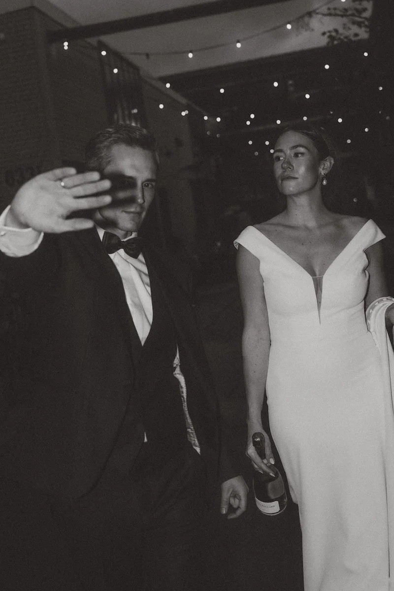 Schwarzweißaufnahme eines Brautpaares, das mit einer Flasche Champagner und einer Hand vor dem Gesicht auf die Kamera zuläuft