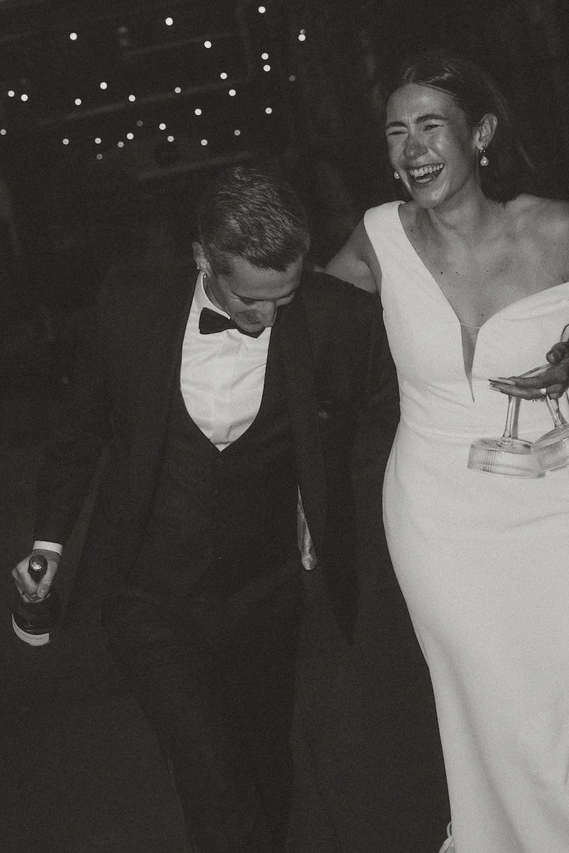 Lachendes Paar, das nach der Hochzeit mit einer Flasche Champagner durch die Straßen von Mallorca läuft