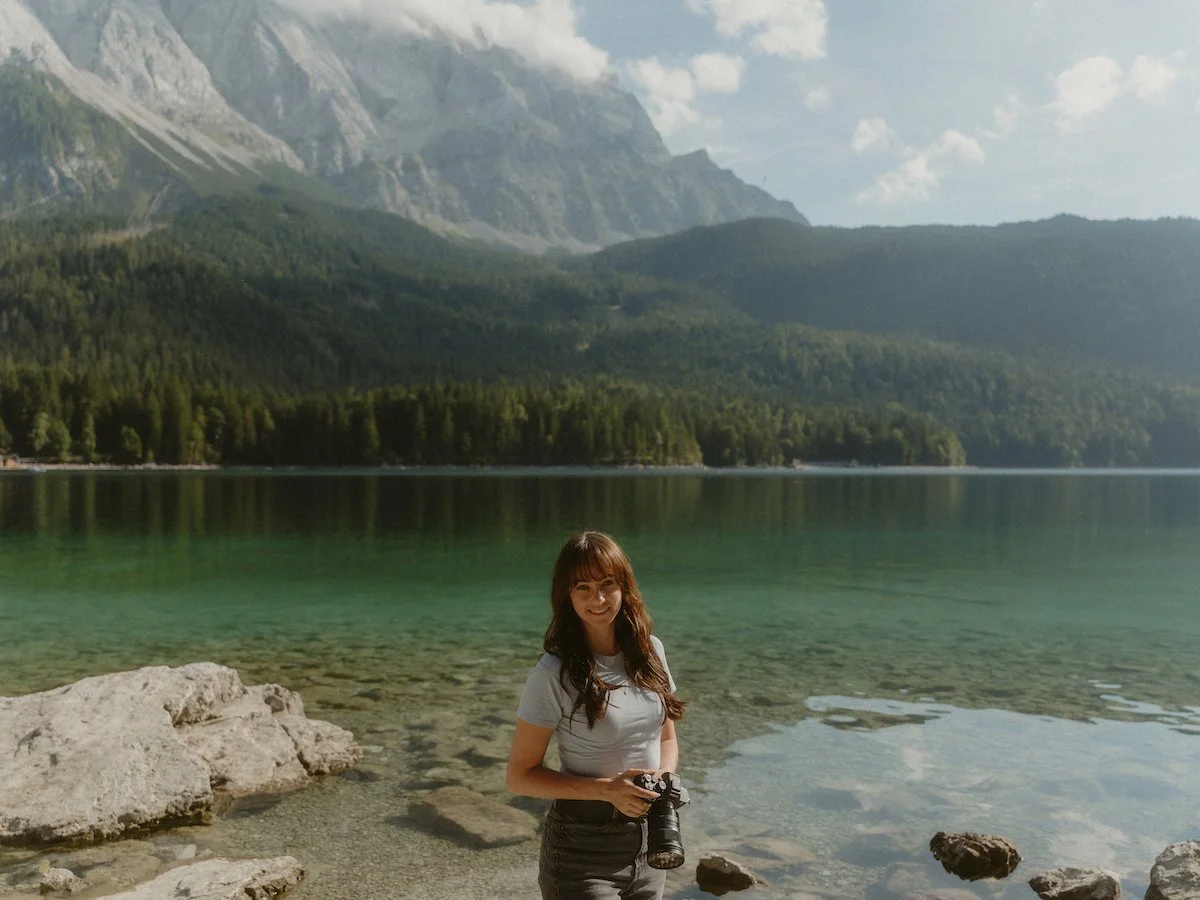 Lächelnde Hochzeitsfotografin mit einer Kamera in der Hand, die vor einem See und einer Bergkulisse in Südtirol steht