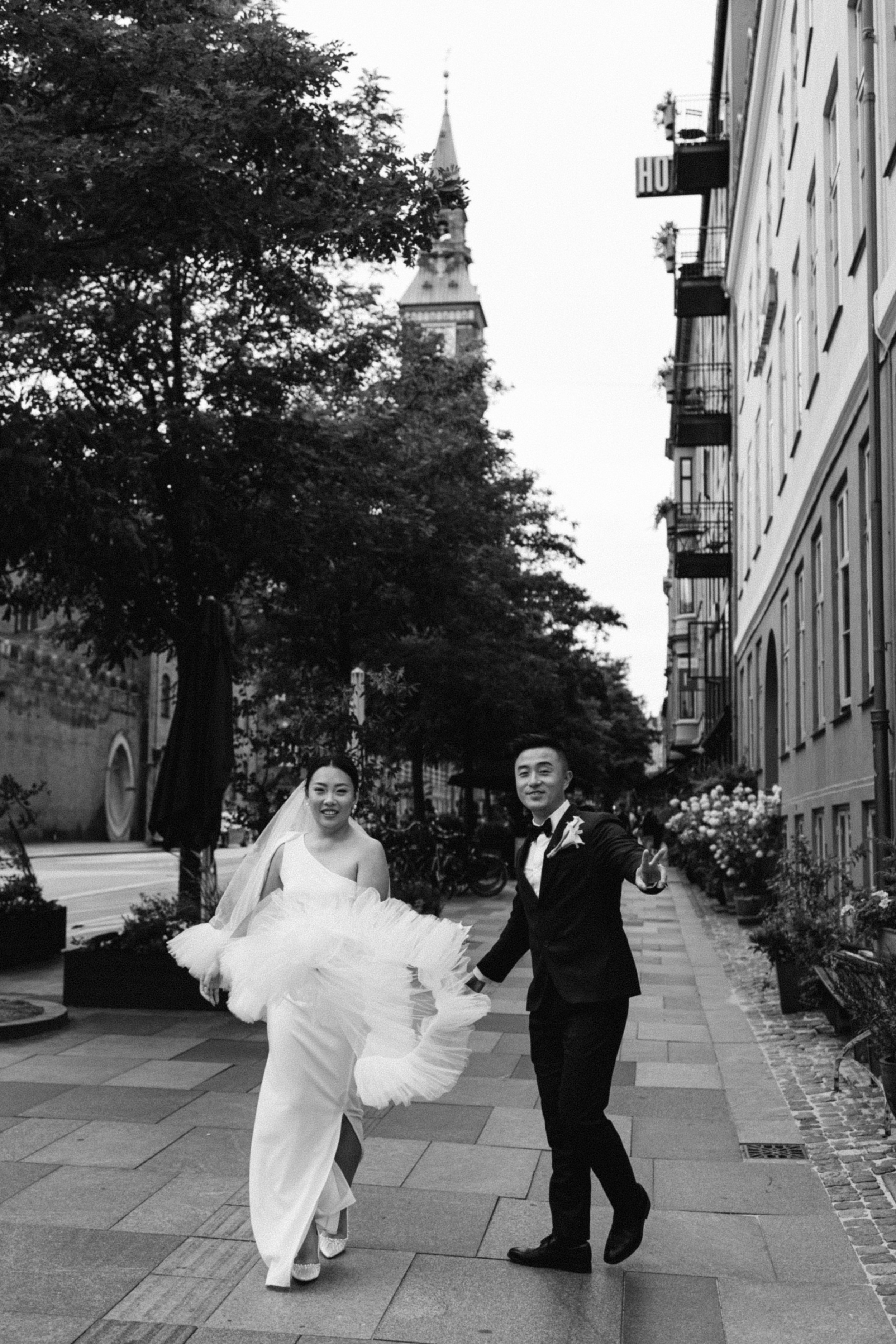A bride and groom walking hand in hand on a city sidewalk, with the bride in a wedding dress and the groom in a tuxedo, smiling and holding hands, with buildings, trees, and a church steeple in the background.