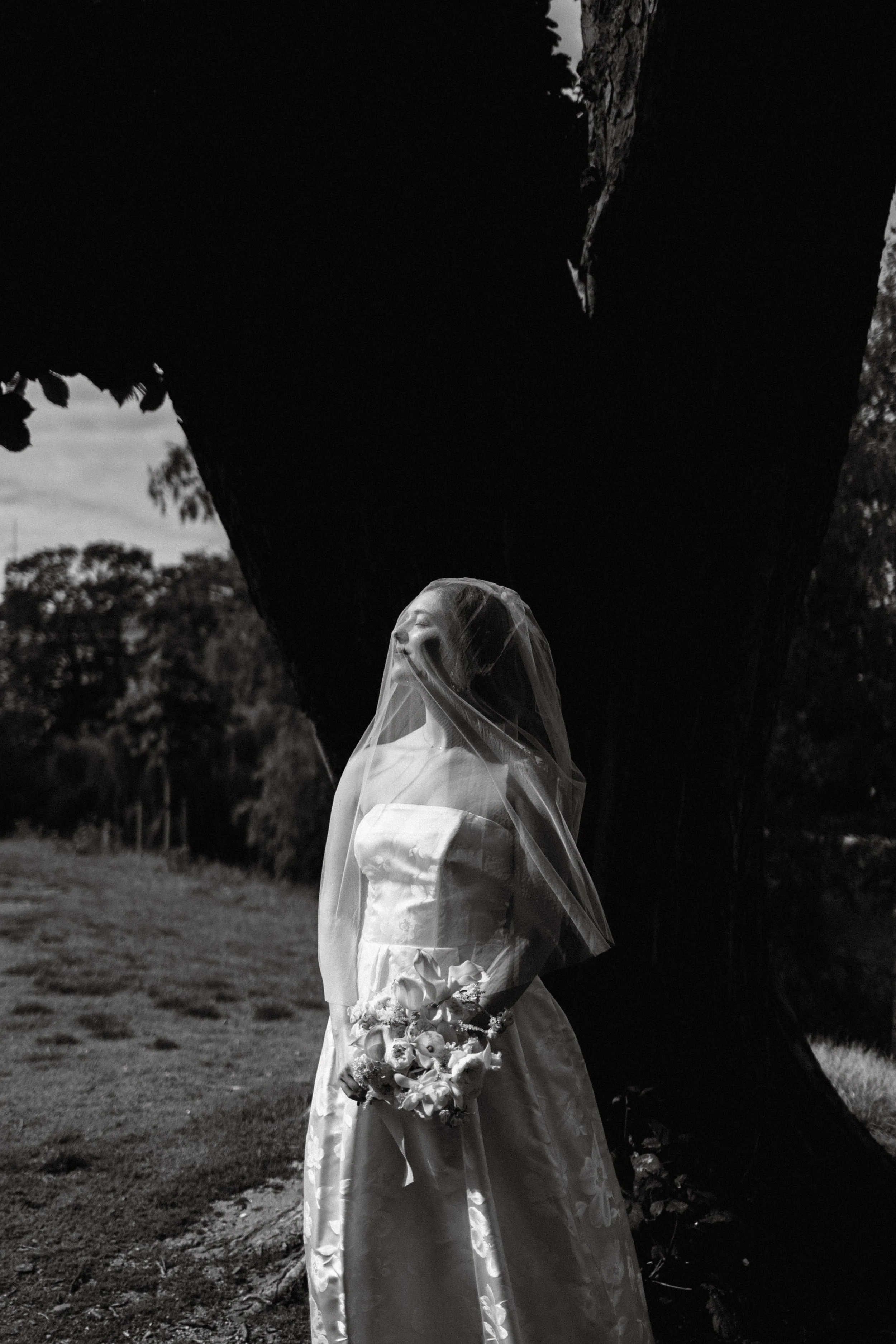A bride in a strapless wedding gown holding a bouquet of flowers, standing outdoors in front of a large, dark tree, with a backdrop of trees and sky, captured in black and white.