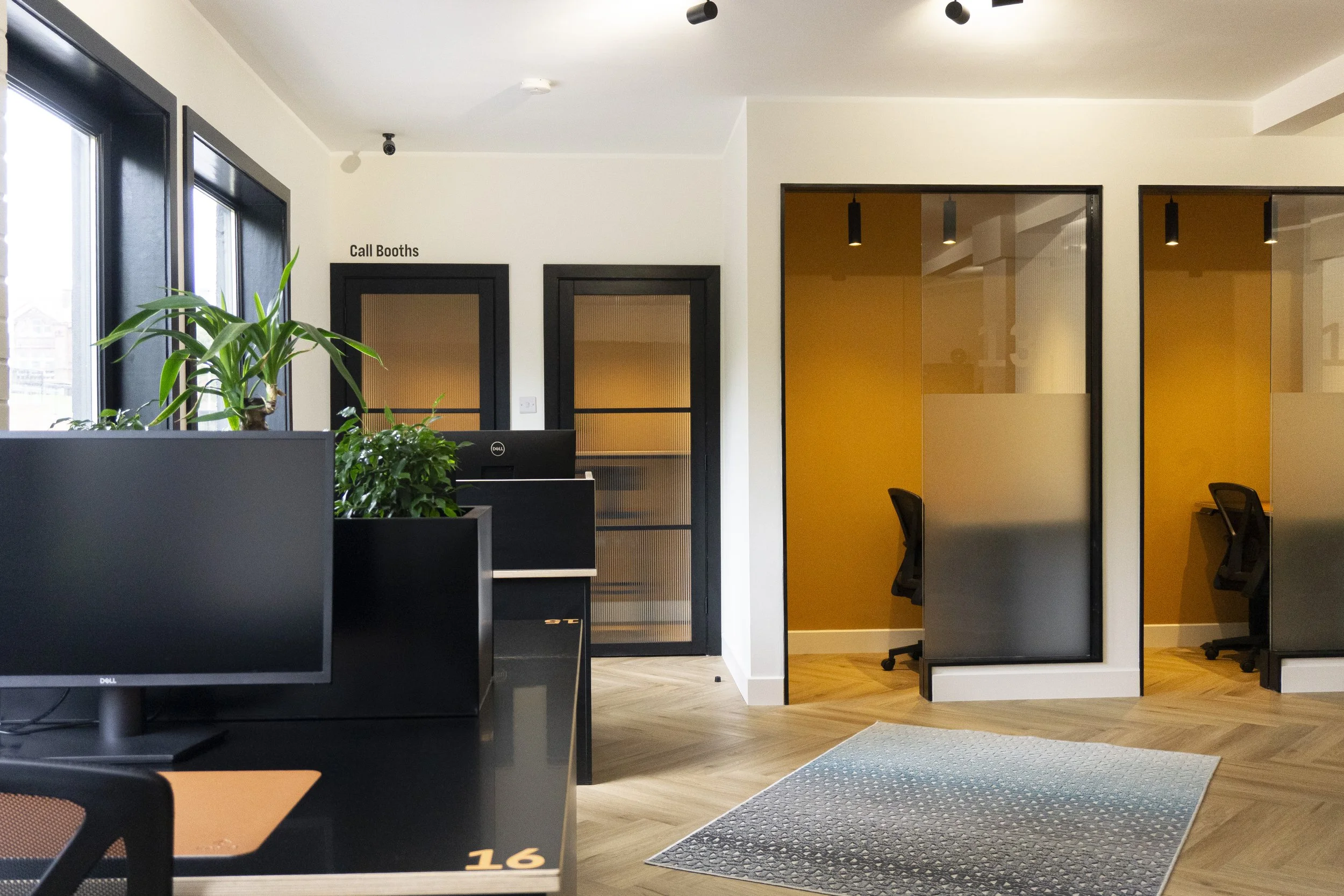 Modern office reception area with black desks, green plants, call booths with black frames, and glass-walled private offices with yellow interior walls.