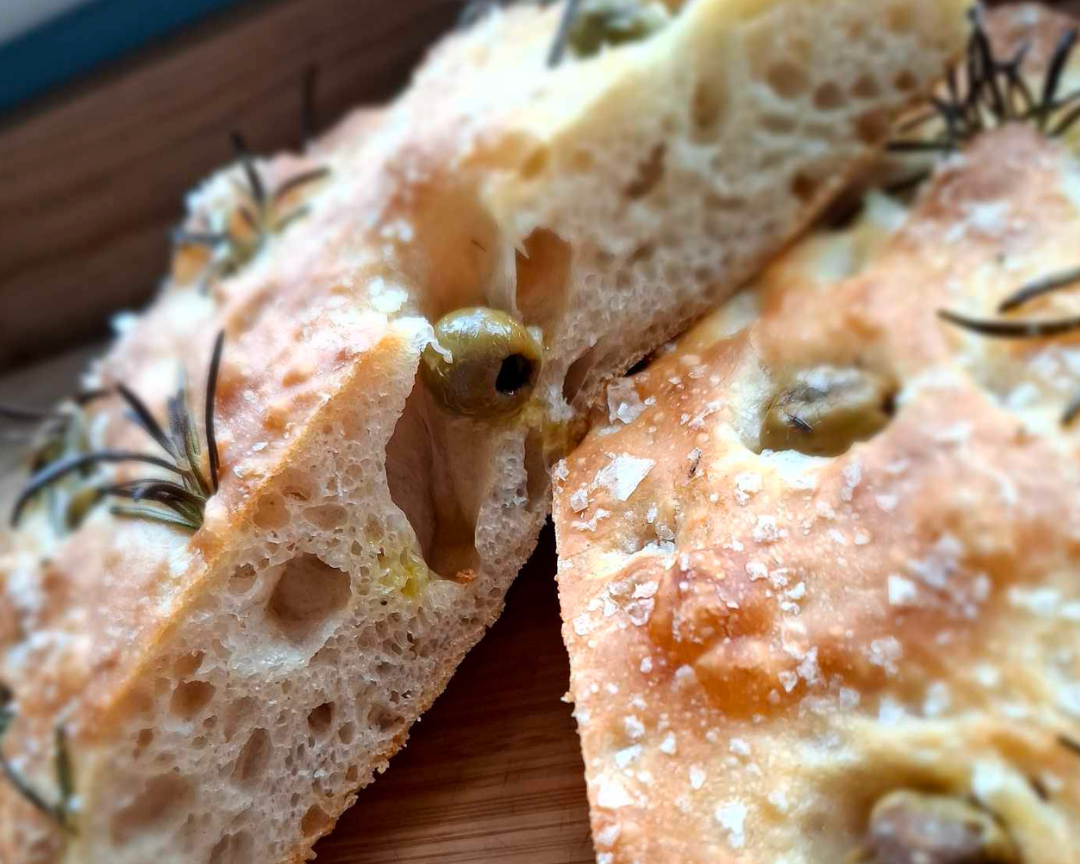 Close-up of a sourdough bread with green olives, rosemary, and sea salt on top, sliced open to show its airy interior.