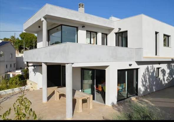 Modern white two-story house with large glass windows and balcony, concrete patio with bench and seating area, overlooking a hillside neighborhood.