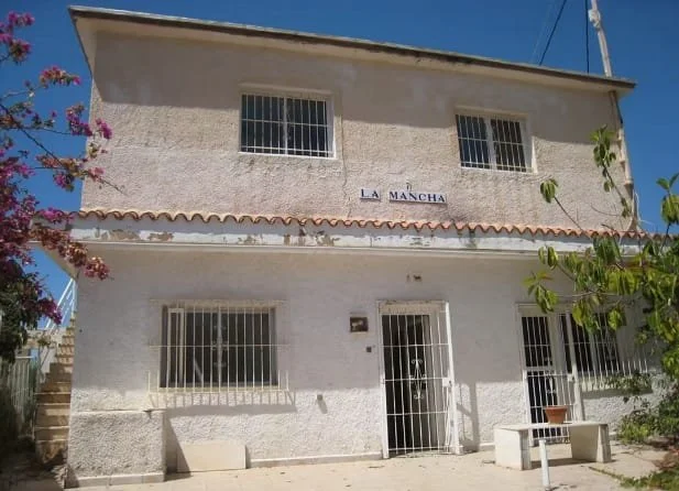 A two-story white house with barred windows and a door, labeled La Mancha, surrounded by plants and flowers, under a clear blue sky.