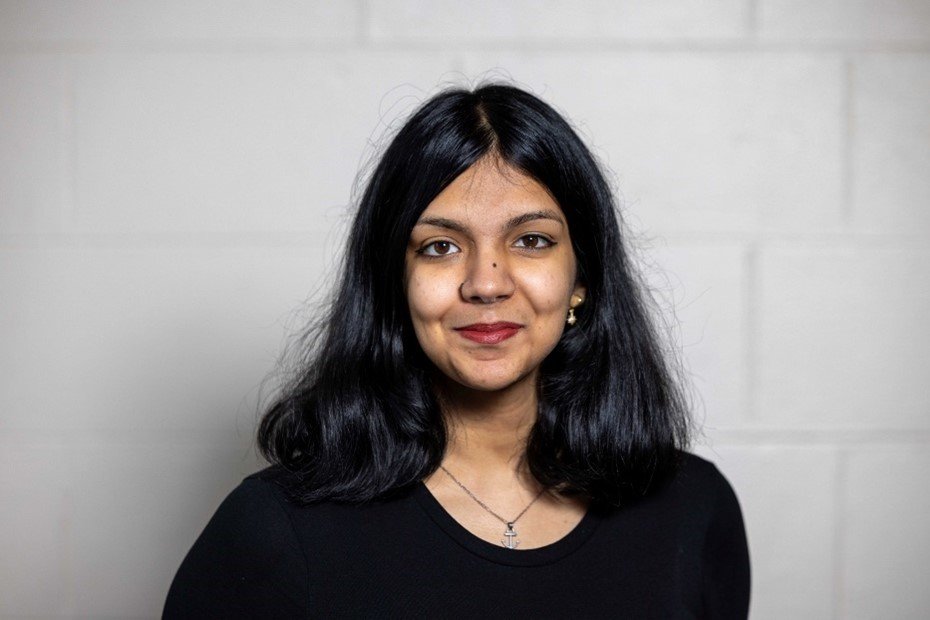 A woman with shoulder-length black hair, wearing a black top and a necklace, standing against a plain light-colored wall.