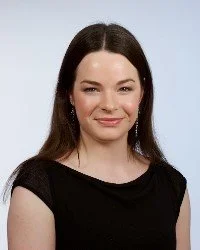 A young woman with long dark hair, wearing a black top, smiling with a light blue background.