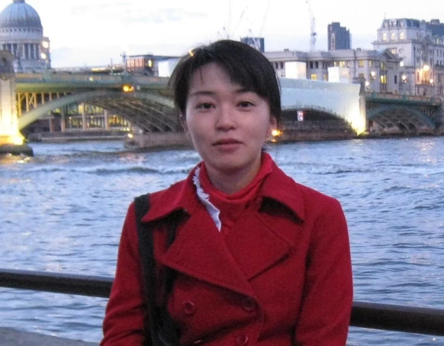 A woman with short dark hair wearing a red coat standing by a river with city buildings and a bridge in the background