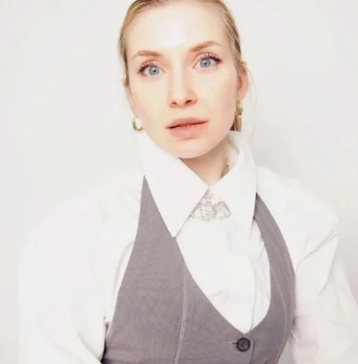 A woman with blonde hair and blue eyes wearing a white shirt and gray vest, posing against a light background.