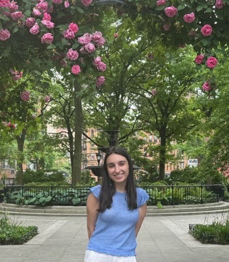 A young woman with long dark hair smiling at the camera, standing on a paved walkway in a park surrounded by green trees and pink flowers above.