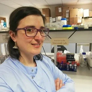A woman in a lab coat with crossed arms in a laboratory, with shelves of supplies and a sign that reads 'Welcome to the Maxwell Lab' in the background.