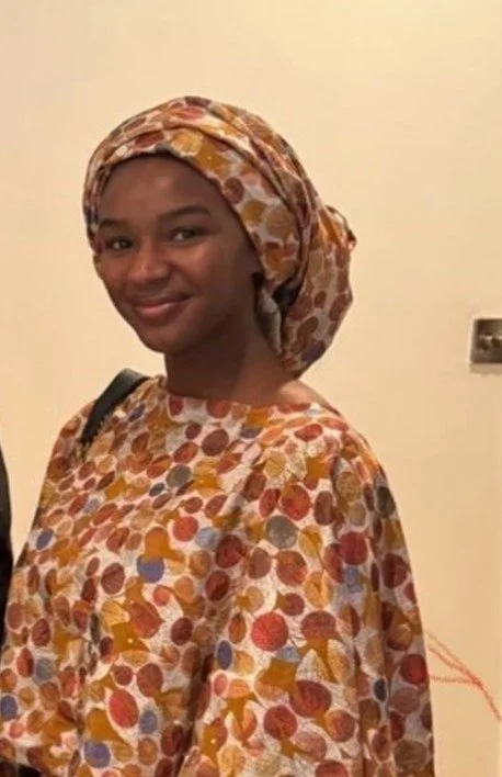 Young woman with dark skin, wearing a patterned headscarf and matching dress, smiling indoors against a beige wall.