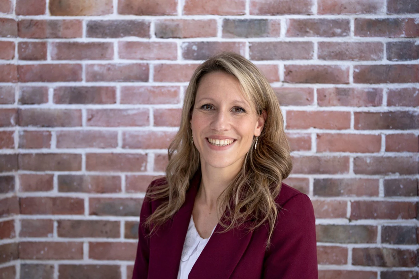 A woman with long blonde hair wearing a burgundy blazer and white shirt, smiling in front of a brick wall.