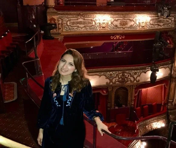 A woman with brown hair smiling and wearing a dark jacket in an ornate theater with red velvet seats and gold detailing.
