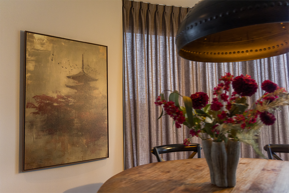 A dining room with a wooden table, a bouquet of pink and red flowers in a gray vase, a large hanging lamp, beige curtains, and a framed painting of a pagoda on the wall.