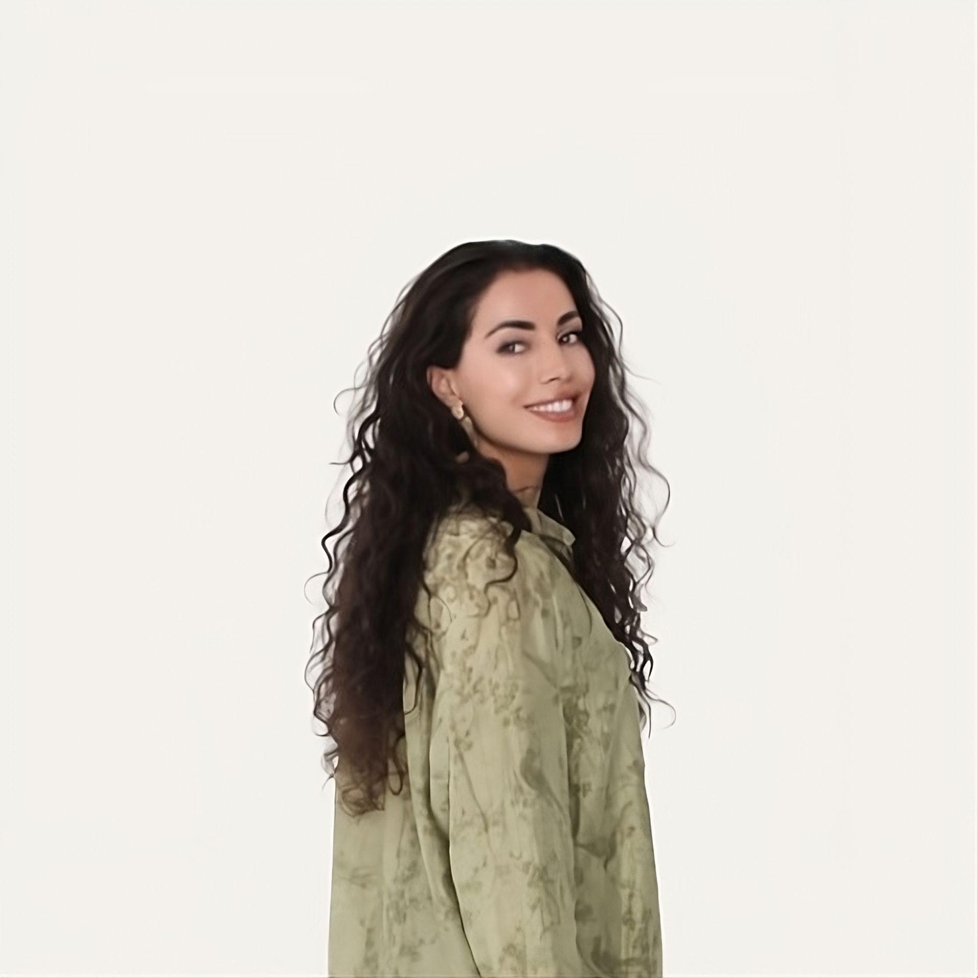 A woman with long, curly dark hair and light skin, wearing a light green patterned blouse and earrings, smiling and looking over her shoulder against a plain white background.
