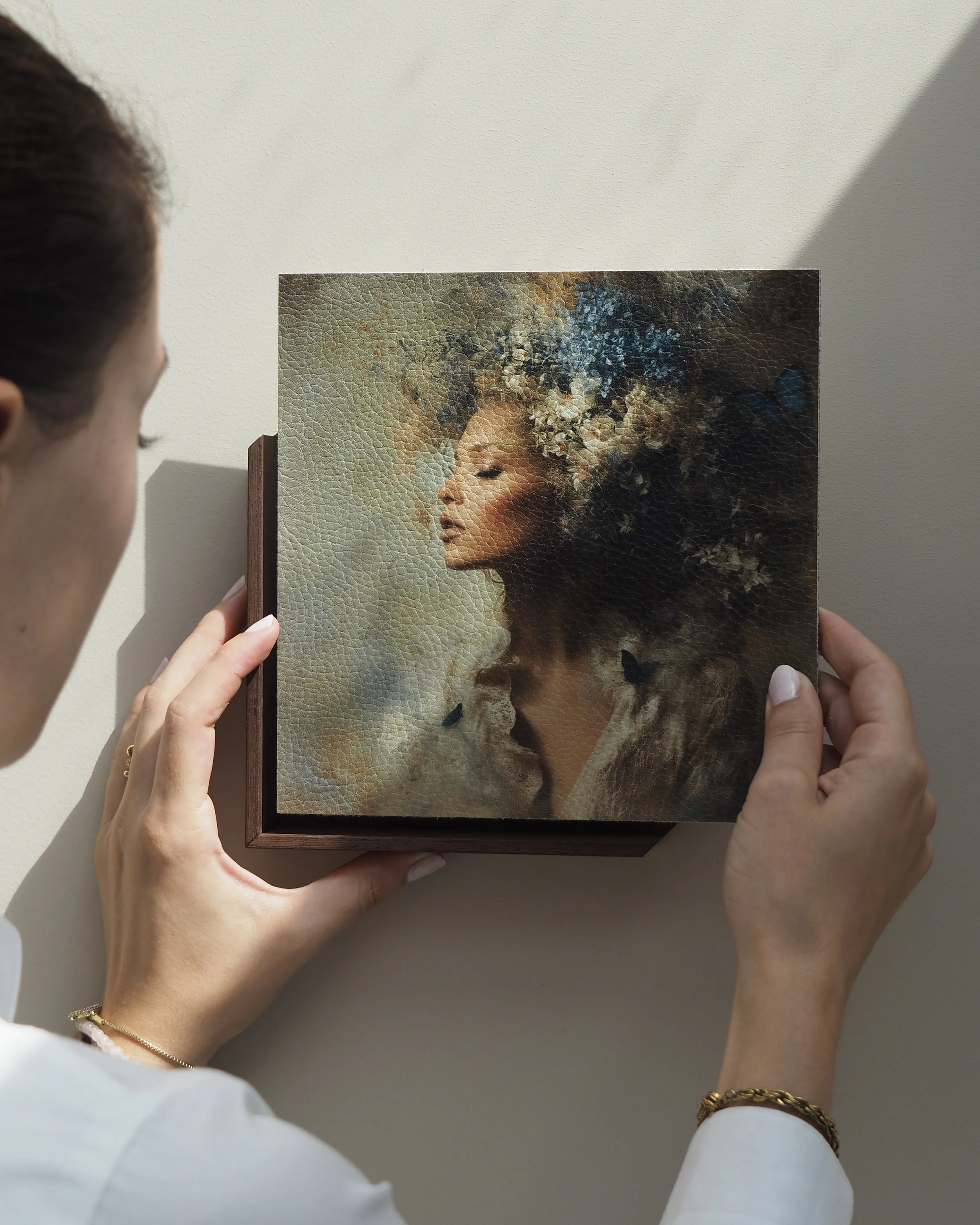 Woman holding a framed painting of a woman with curly hair adorned with flowers, with a dark and blue butterfly nearby.