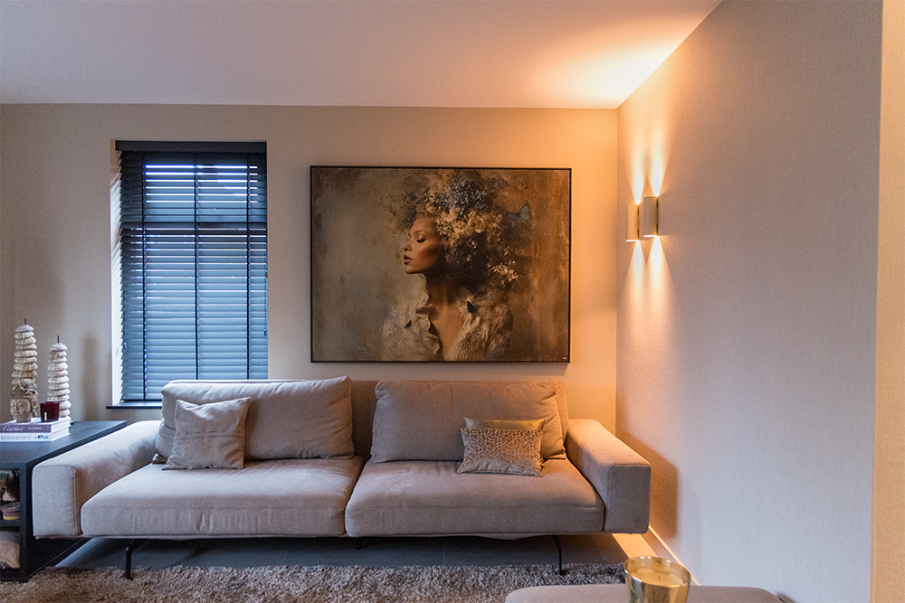 Modern living room with a beige sofa, decorative pillows, a painting of a woman with floral hair, lamp on the right wall, window with blinds on the left, and small decor items on the side table.