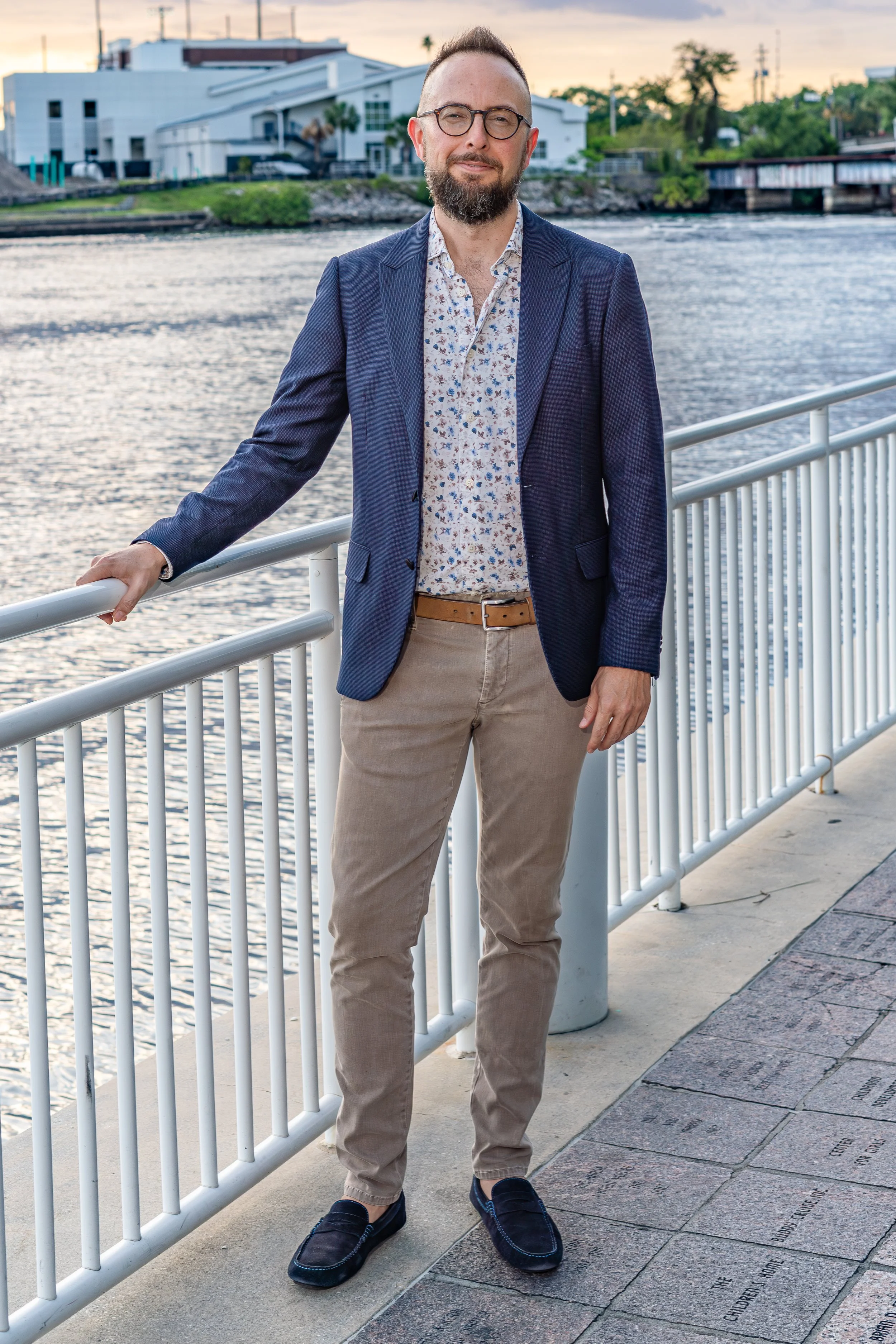 A man with glasses and a beard standing outdoors by a river, wearing a dark blue blazer, floral shirt, beige pants, and loafers, with a cityscape and sunset in the background.