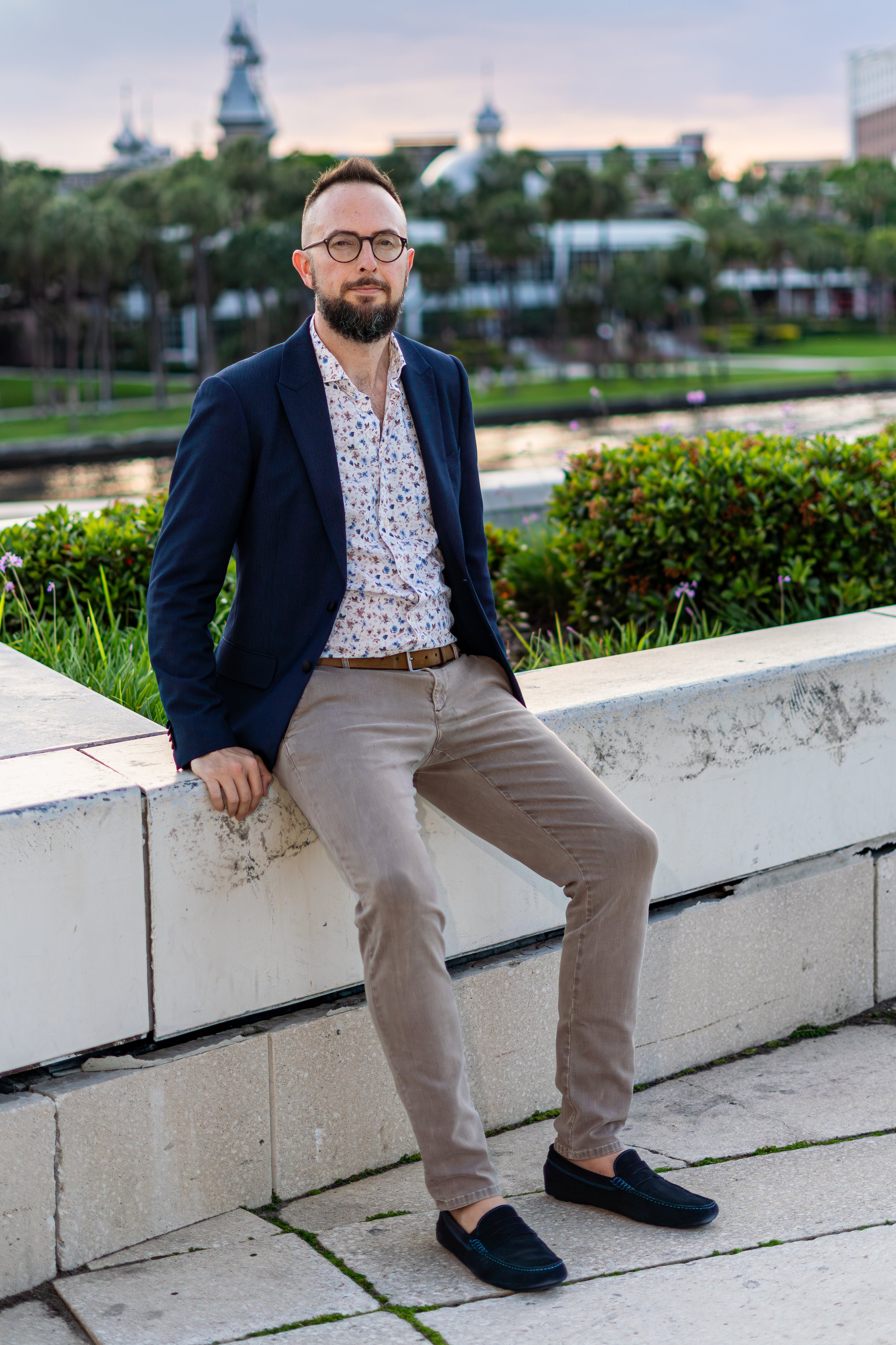 A man with glasses and a beard sitting on a white concrete ledge outdoors, with a cityscape and water in the background.