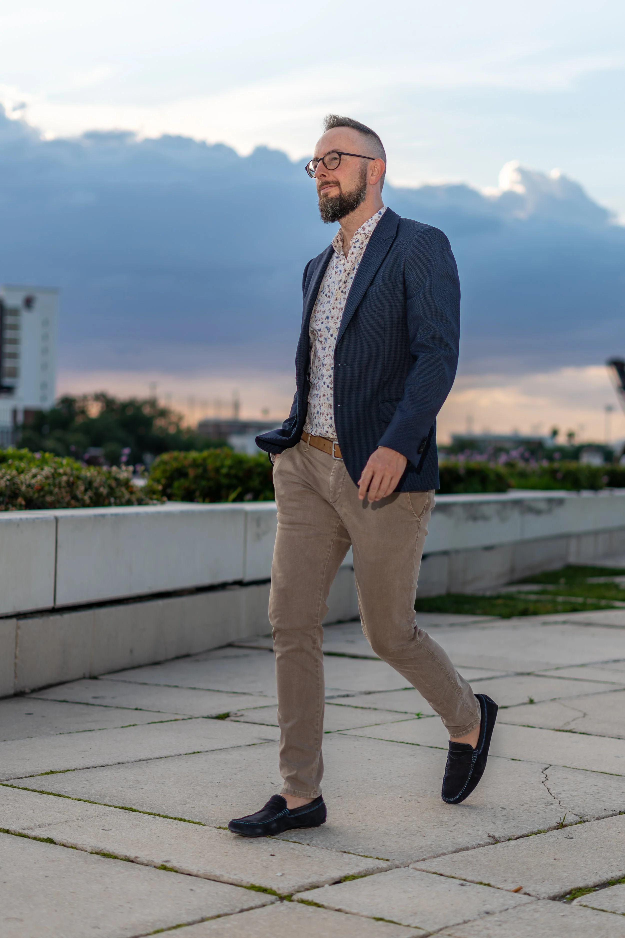 A man in a blue blazer, patterned shirt, beige pants, and black shoes walking outdoors on a paved area with a cityscape and cloudy sky in the background.