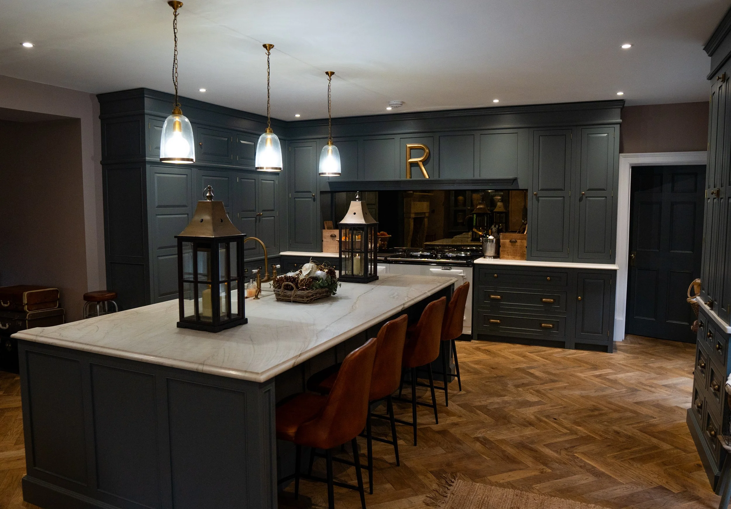 Modern kitchen with dark gray cabinets, a large white marble island, three pendant lights above, and hardwood flooring. Decor includes lanterns, a basket, and a gold letter R on the wall.