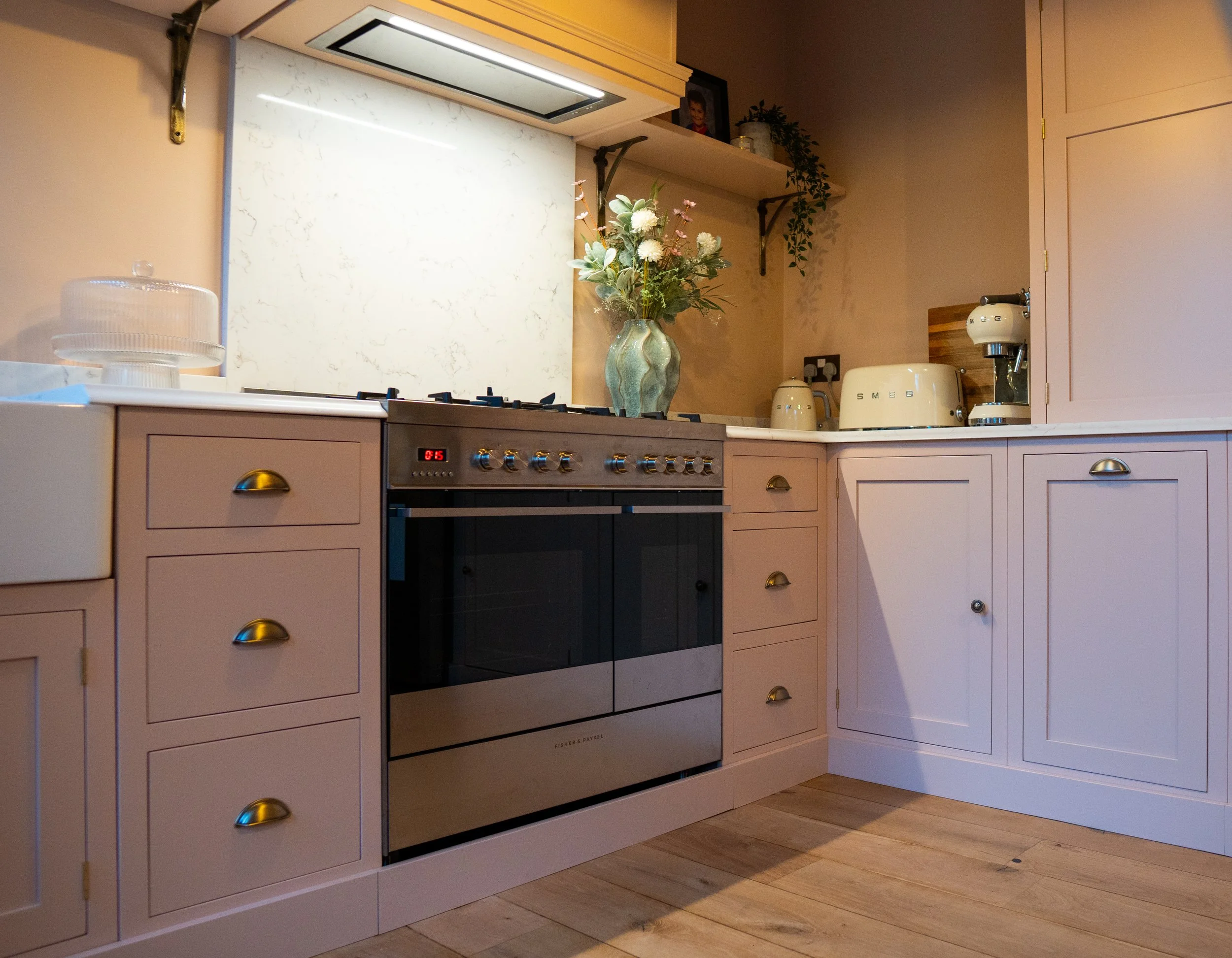 A kitchen with light pink cabinets, a black oven, a vase with white and pink flowers, and various small appliances on the countertop.