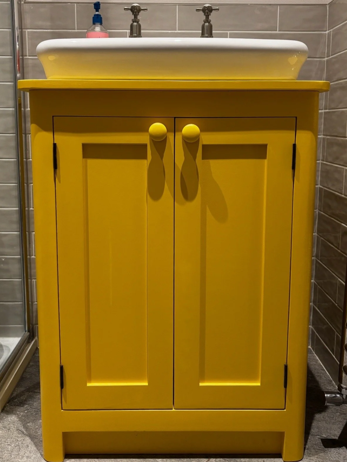 Bright yellow bathroom vanity with two cabinet doors, each with round yellow knobs, and a white sink on top. A soap dispenser is visible on the left side of the sink. The background features gray tiled walls.