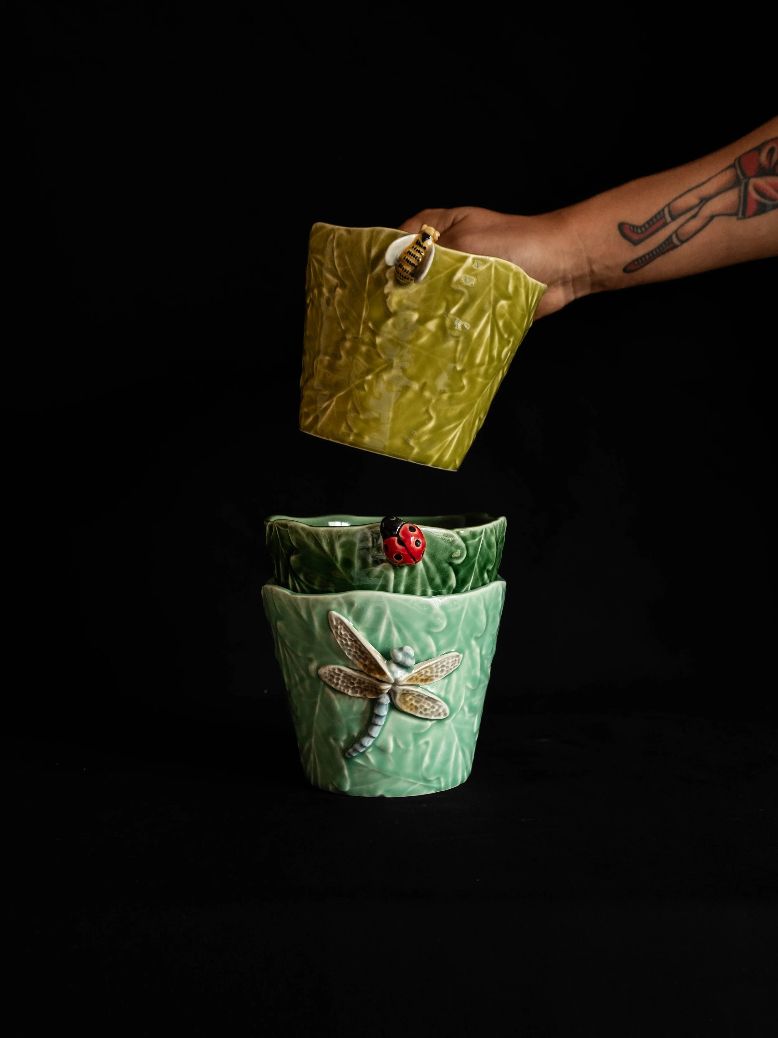 A person's arm with a tattoo holding a yellow ceramic container above two green ceramic containers, each decorated with an insect ornament, against a black background.