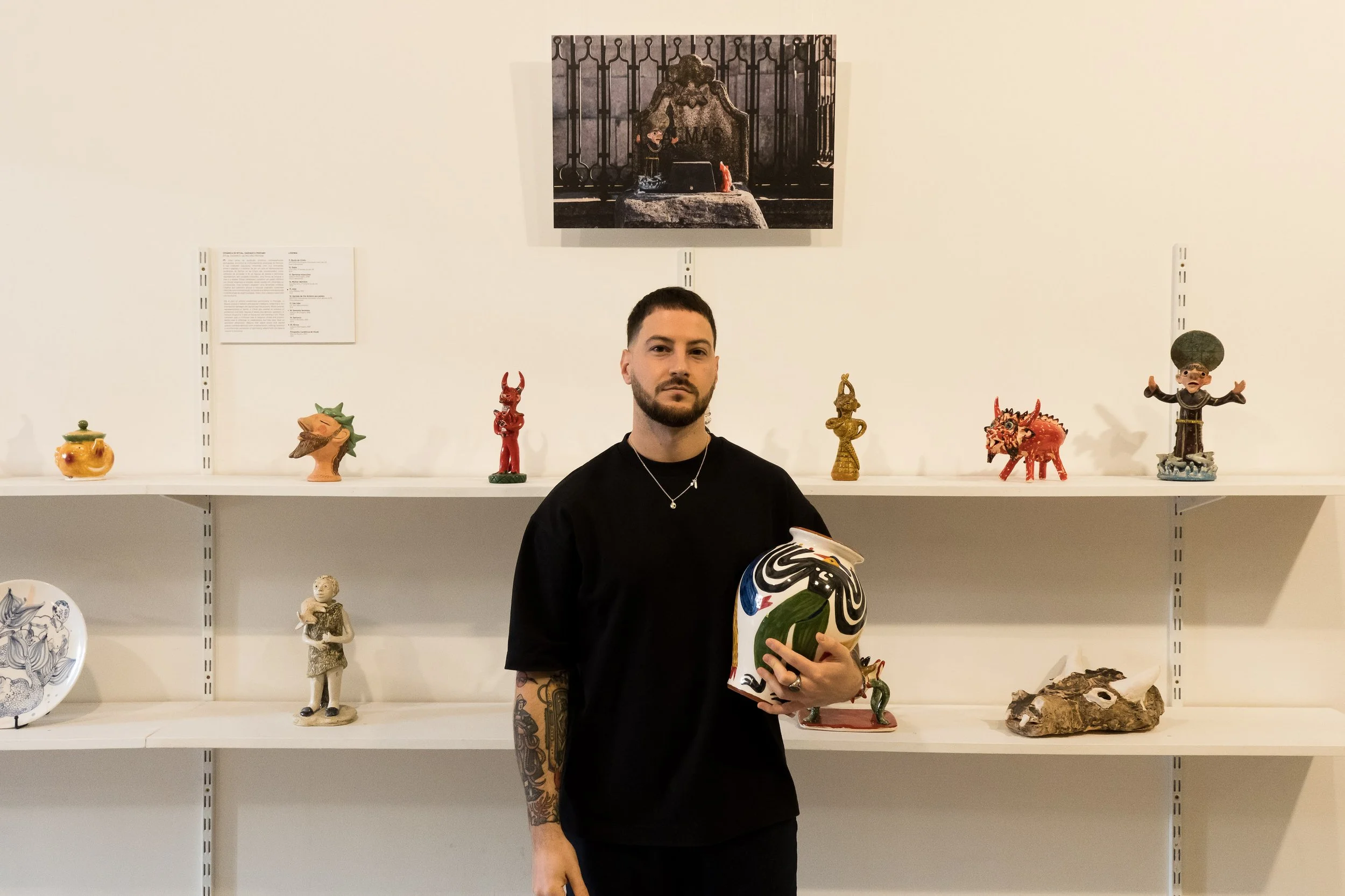 A man with short dark hair, beard, and tattoos on his left arm stands in an art gallery, holding a decorative ceramic piece with bold black, white, red, and green designs. He wears a black t-shirt and a silver necklace. Behind him, a white wall and shelves display colorful ceramic and sculptural art pieces. A framed black-and-white photograph of a monument or sculpture is on the wall above him.
