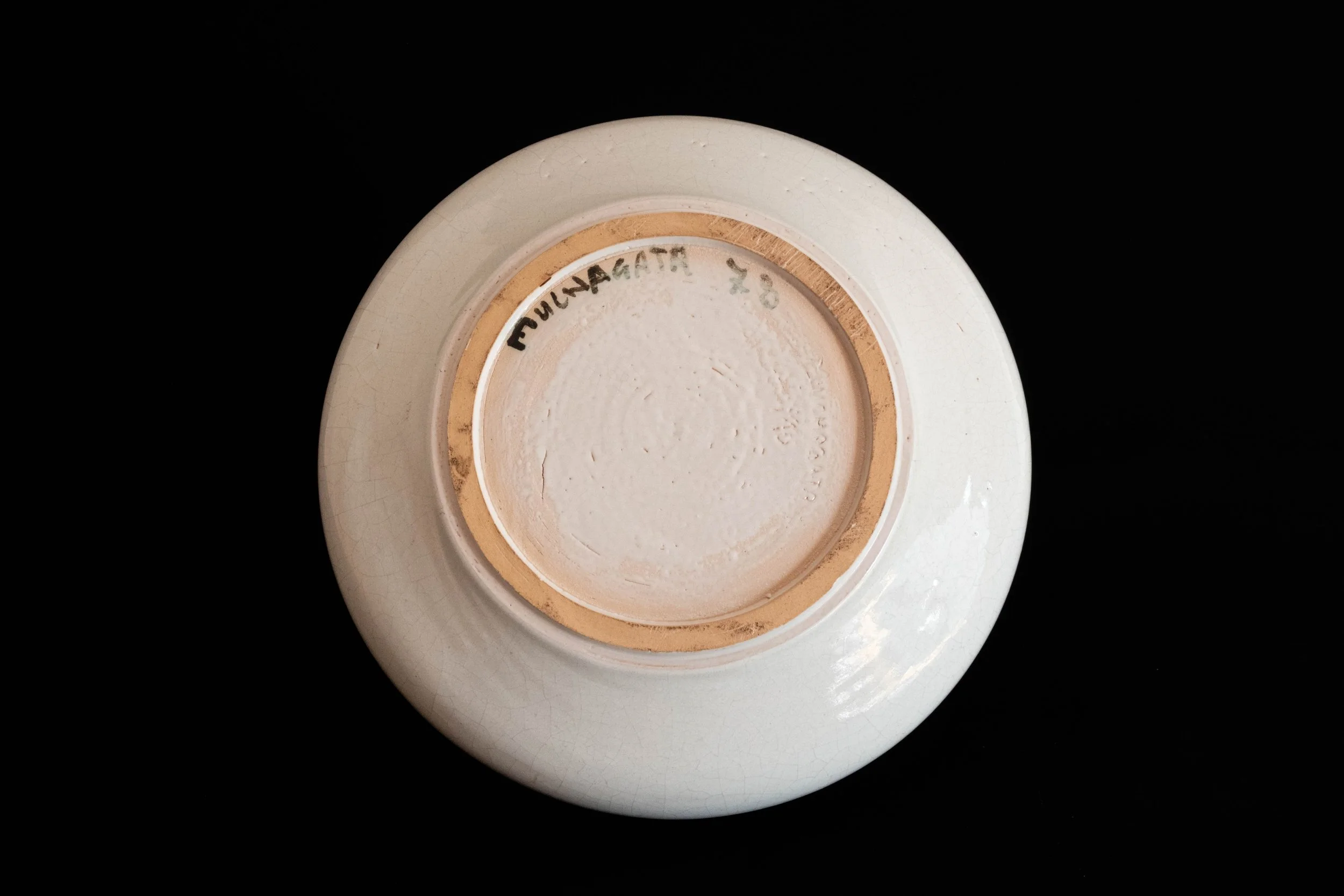Underside of a white ceramic cup with a gold-colored rim, with handwritten text 'Fumagatte 58' on the bottom, against a black background.