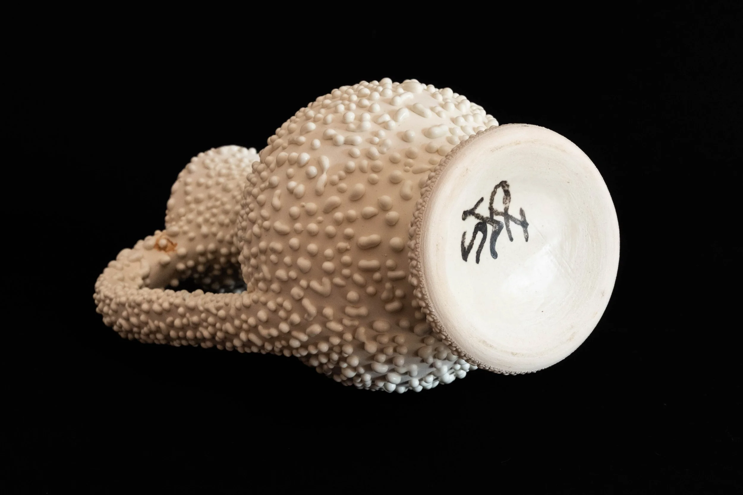 A ceramic sculpture of a human skull covered in small, rounded protrusions, lying on its side against a black background, with a painted face on the underside visible.