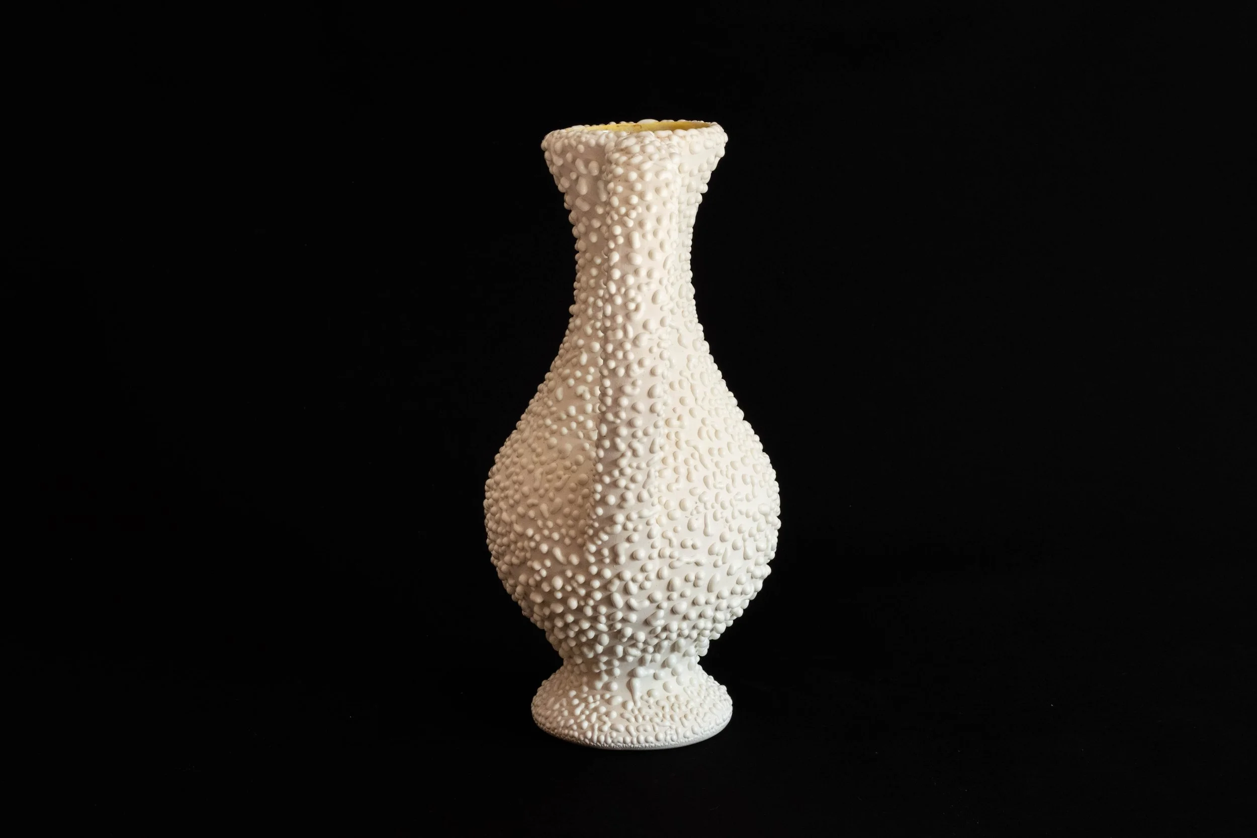 A white ceramic vase with a textured surface covered in small, rounded protrusions, standing on a black background.