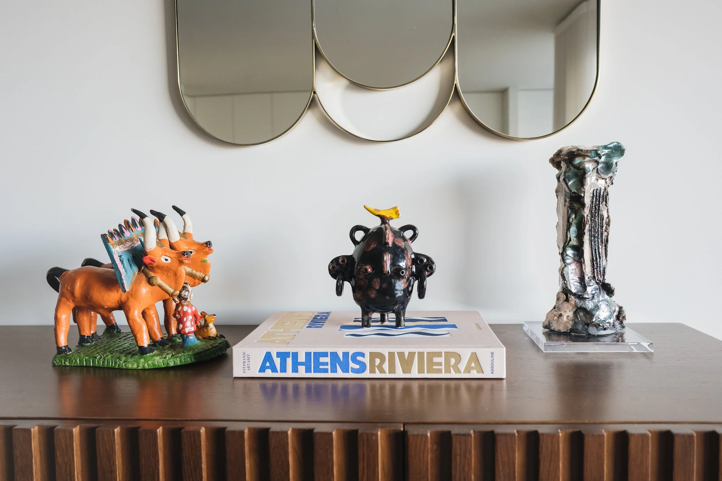 Decorative figurines on a wooden table, including a black cow with a yellow banana on its head, an orange herd of cattle with black horns, and a tall abstract sculpture with metallic textures. There's a book titled 'ATHENS RIVIERA' under the cow figurine. A modern mirror with rounded sections is hung on the white wall in the background.