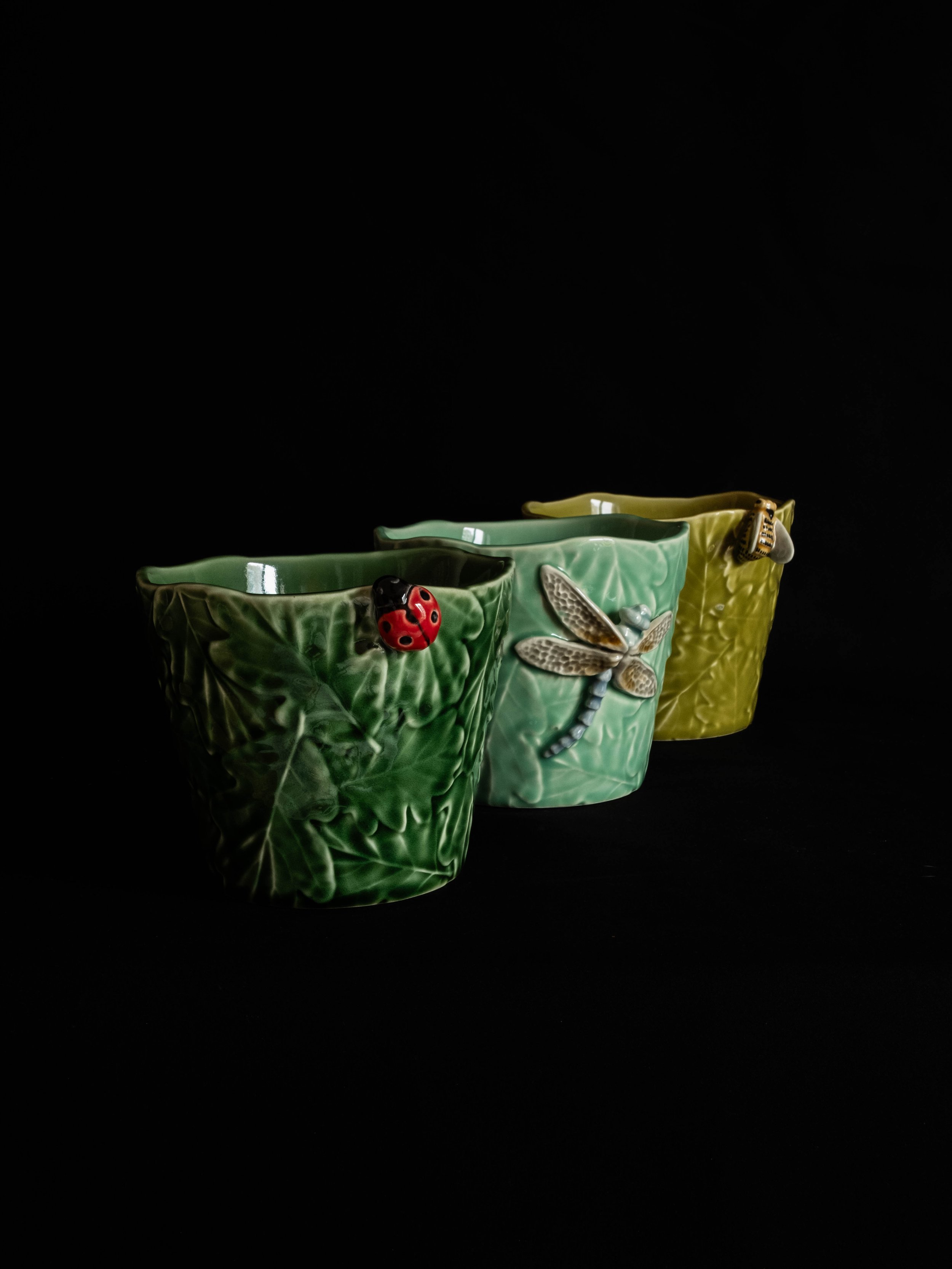 Three ceramic cups with colorful insect designs, including a ladybug, a dragonfly, and a bee, set against a black background.